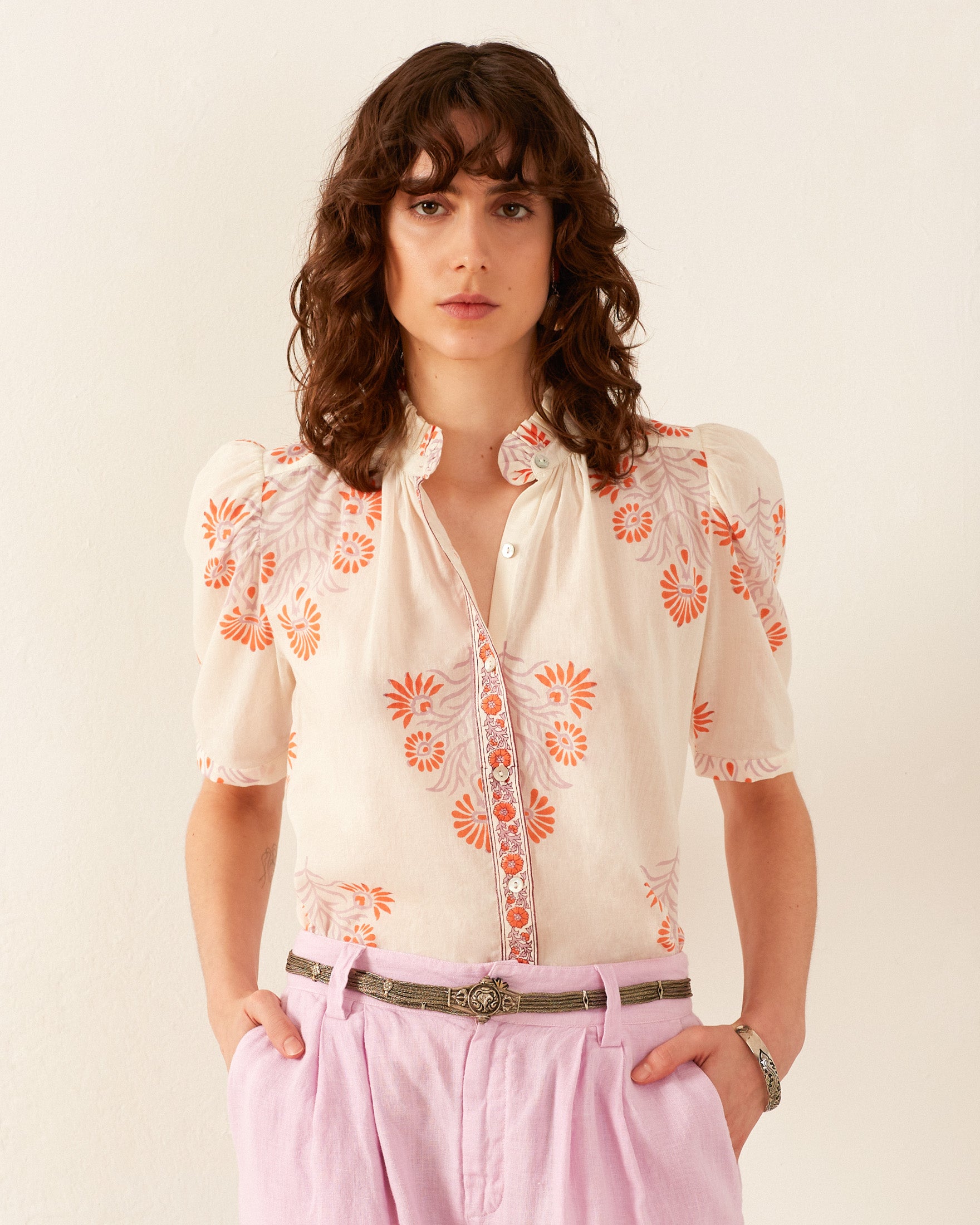 Someone with curly hair is wearing a Winn Papaya Aster Shirt, designed with vintage-inspired orange floral patterns, and they have teamed it up with lavender pants. They are positioned against a plain background, gazing at the camera with a neutral expression.