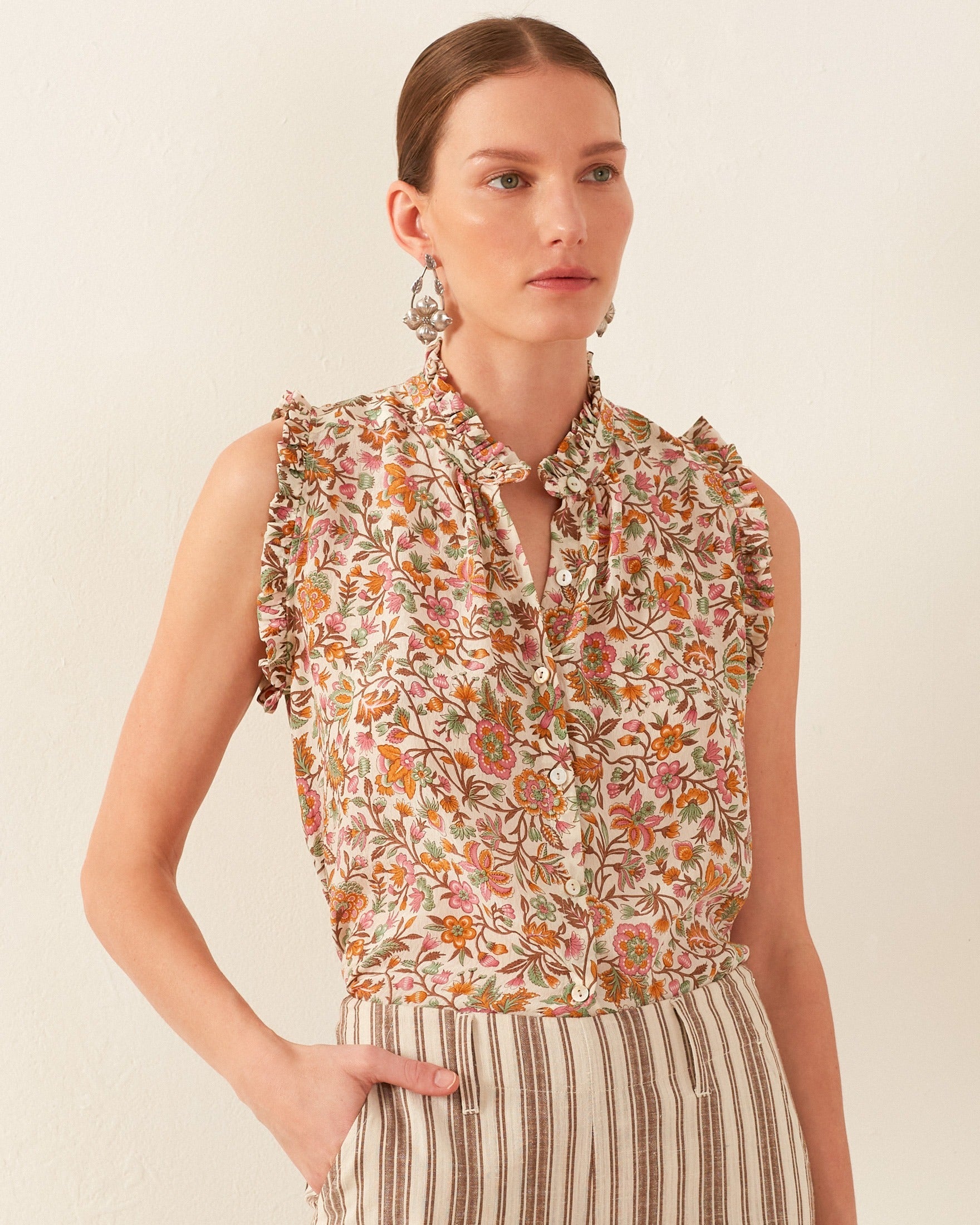 A person with tied-back hair wears a vintage-inspired sleeveless blouse with ruffled details and striped pants, complemented by the geometric print of the Wylder Garden Party Silk Shirt peeking through. Standing against a plain light background, they casually place one hand in their pocket.