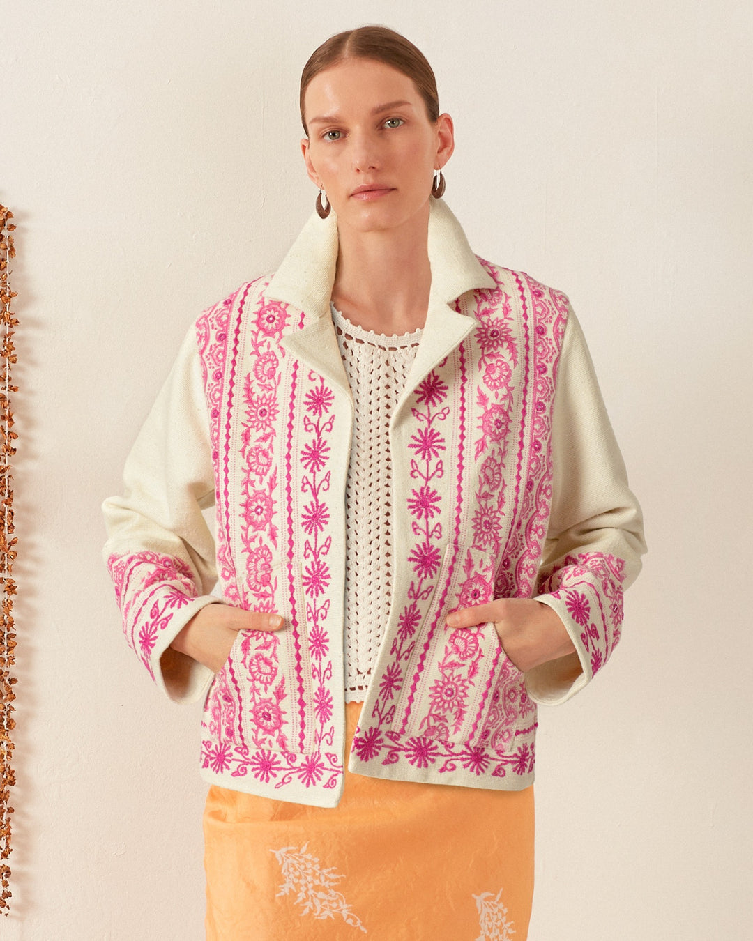 A person stands against a cream-colored wall, wearing the Western Phlox Riviera Jacket, which features pink floral hand embroidery over a knitted cream top and an orange skirt adorned with white flowers. They have their hands casually tucked into the jacket pockets.