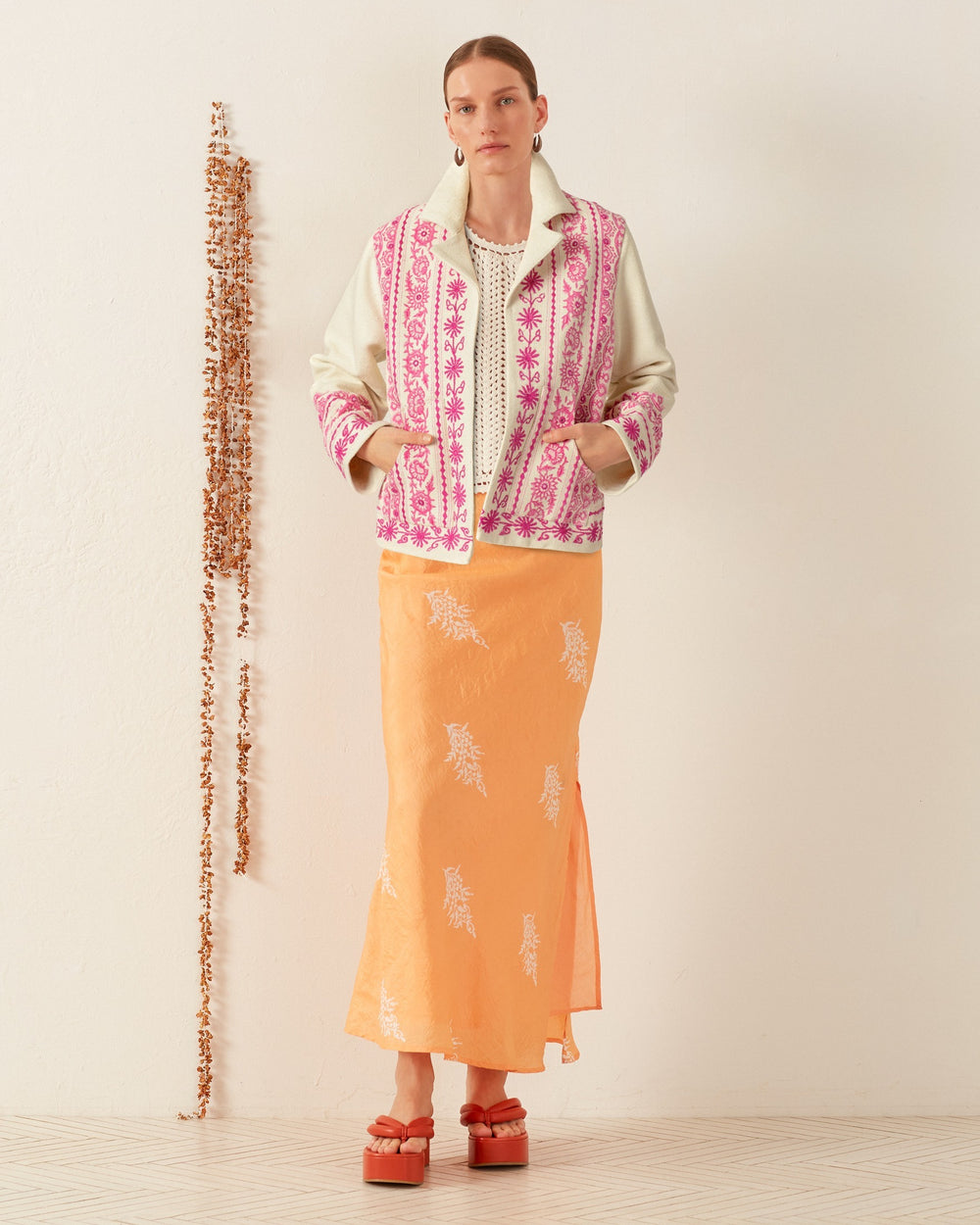 A person stands against a cream wall wearing the Western Phlox Riviera Jacket, which is white and pink with hand embroidery. They pair it with an orange skirt featuring white patterns and red platform sandals. A vertical row of small decorative items is on the wall beside them.