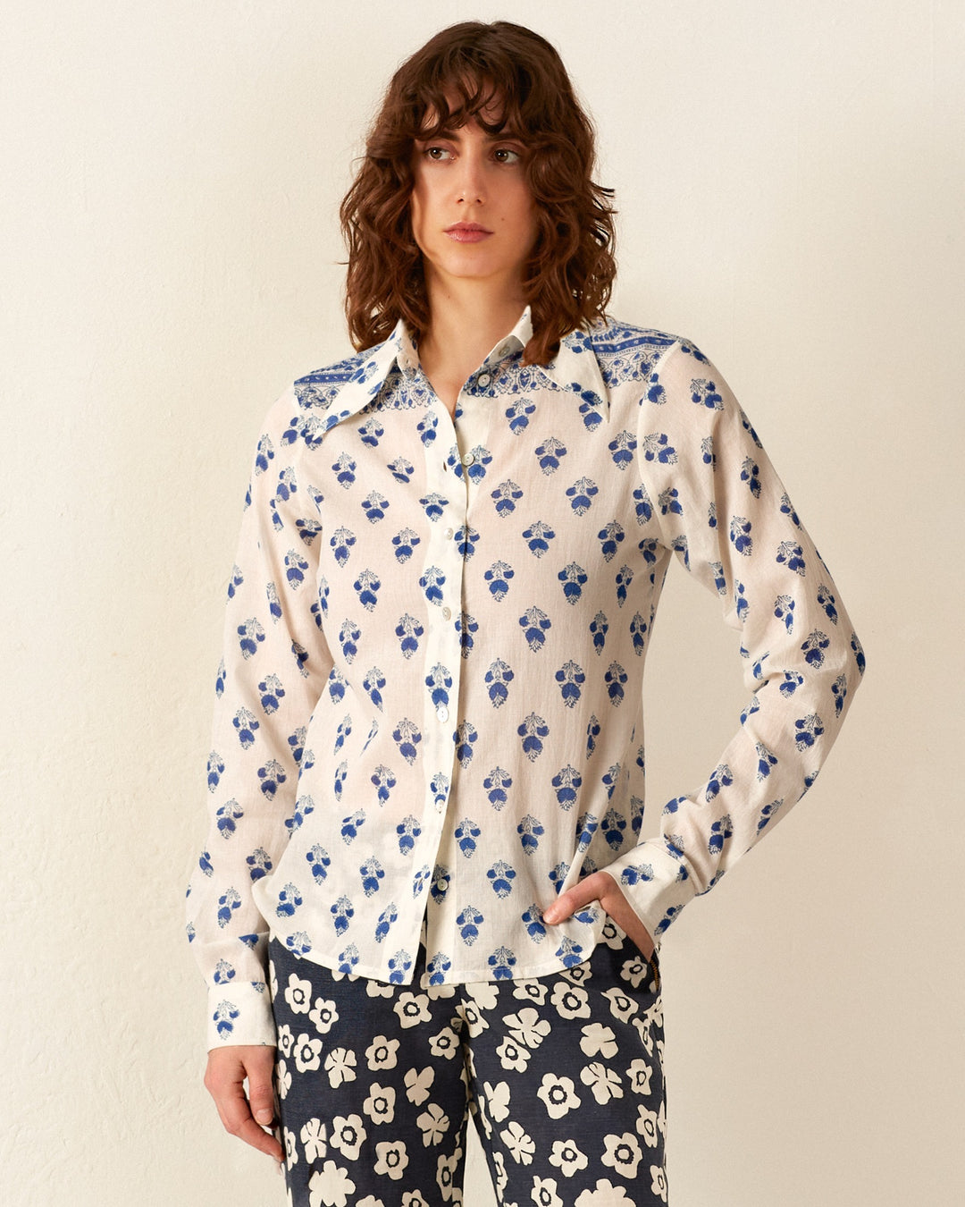 A person with curly hair is wearing the Dolly Indigo Bhutti Shirt, a white button-up featuring blue floral patterns, and black high-waisted pants adorned with hand-block printed white floral designs. They are standing against a plain white wall.