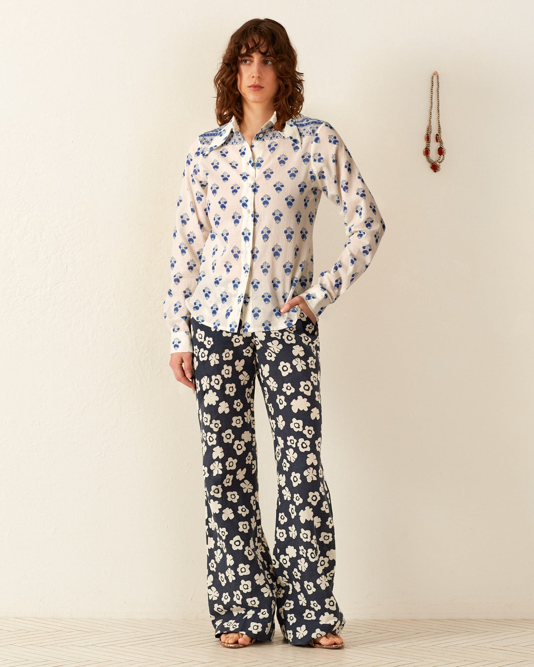 A person with medium-length wavy hair stands against a plain wall, wearing the Dolly Indigo Bhutti Shirt and high-waisted pants featuring hand-block printed white floral designs. A necklace hangs on the wall nearby.