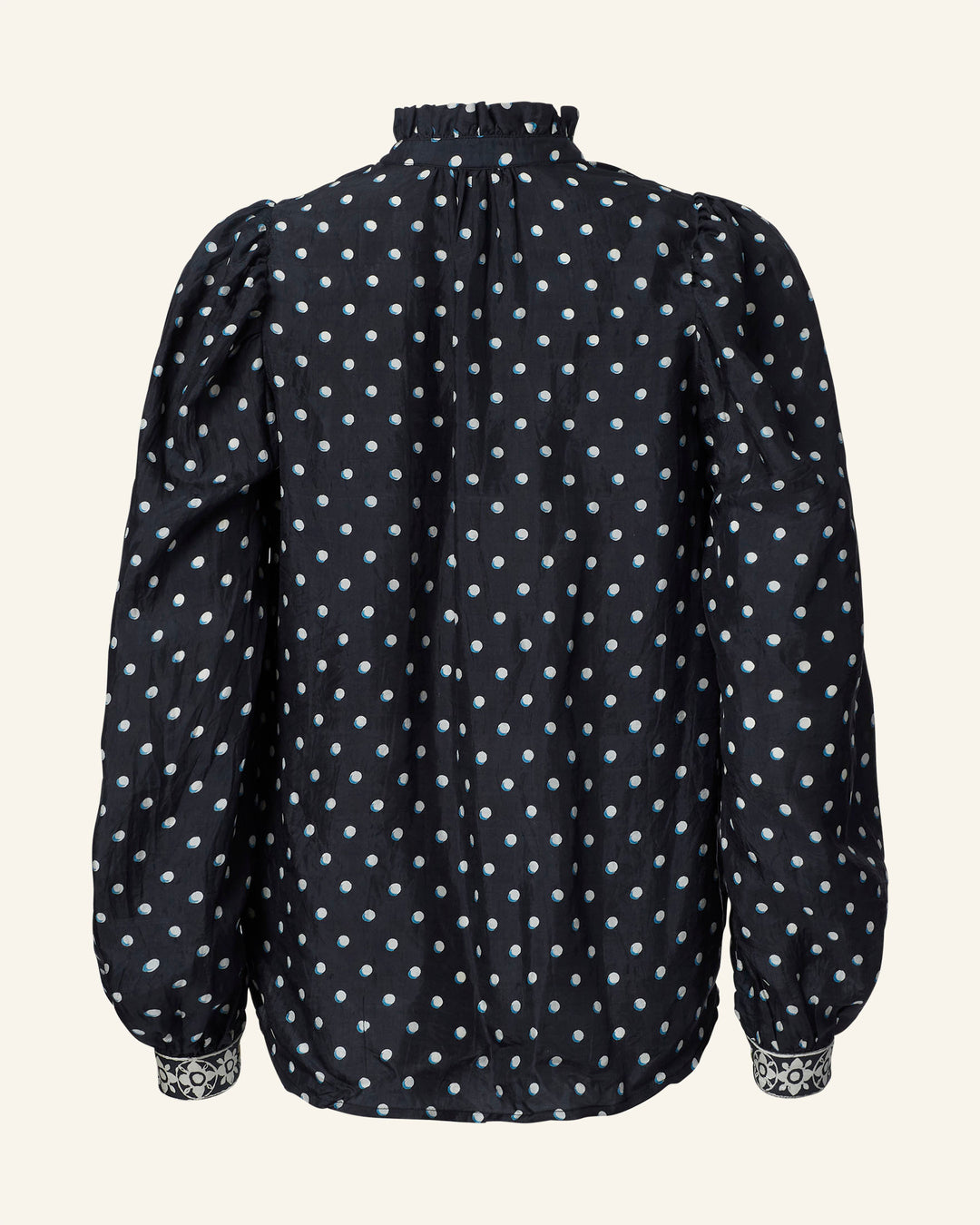 The Annabel Raven Dot Silk Shirt is a black long-sleeve blouse adorned with white polka dots and features ruffled detailing on the collar and cuffs. It showcases puffed shoulders, offers a loose fit, and has contrasting white and black patterned trim on the cuffs, all set against a plain beige background.