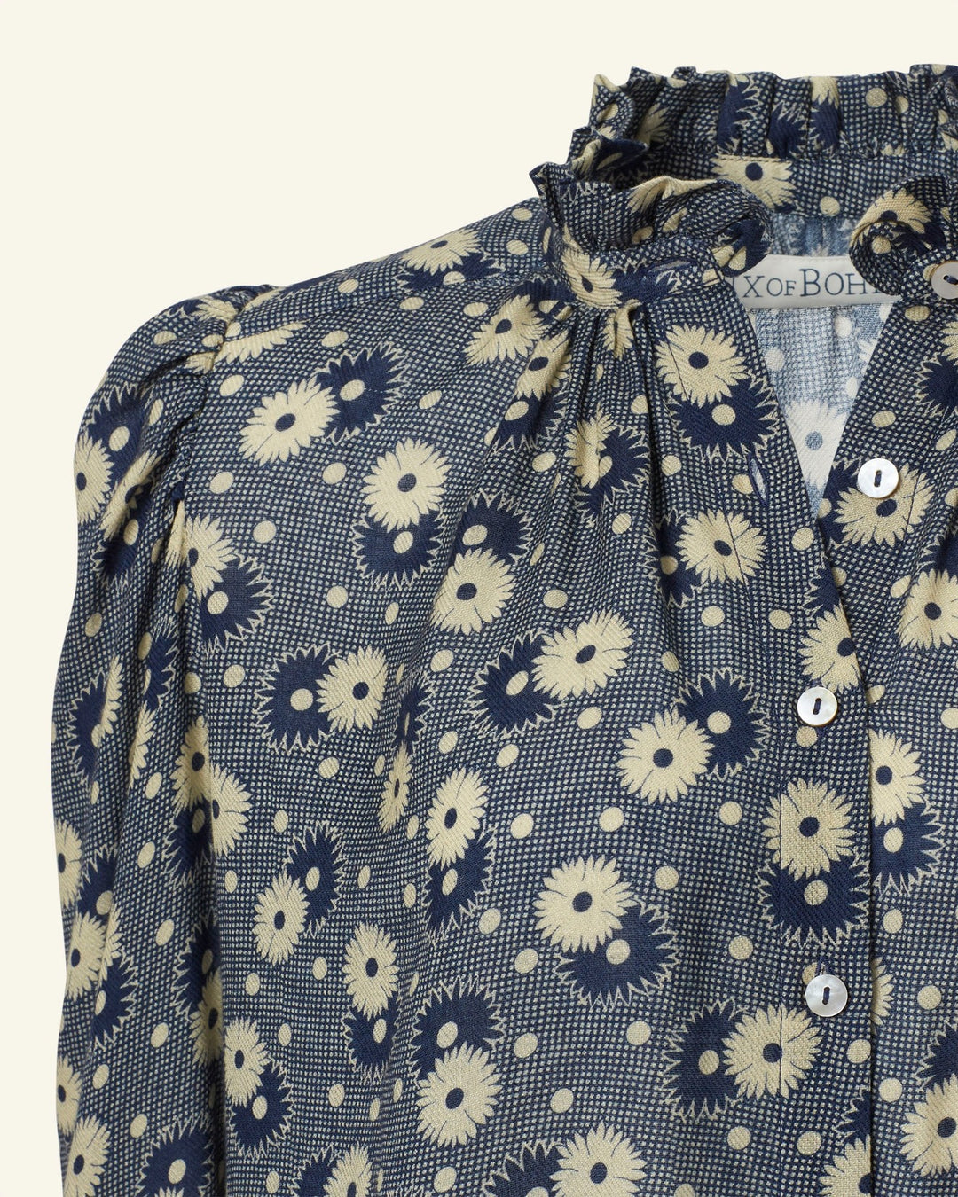 The Artemis Navy Daisy Shirt showcases a close-up of a navy base adorned with a light yellow '70s floral print. The blouse is designed with a ruffled high collar and button-down front, capturing the essence of vintage-inspired charm.