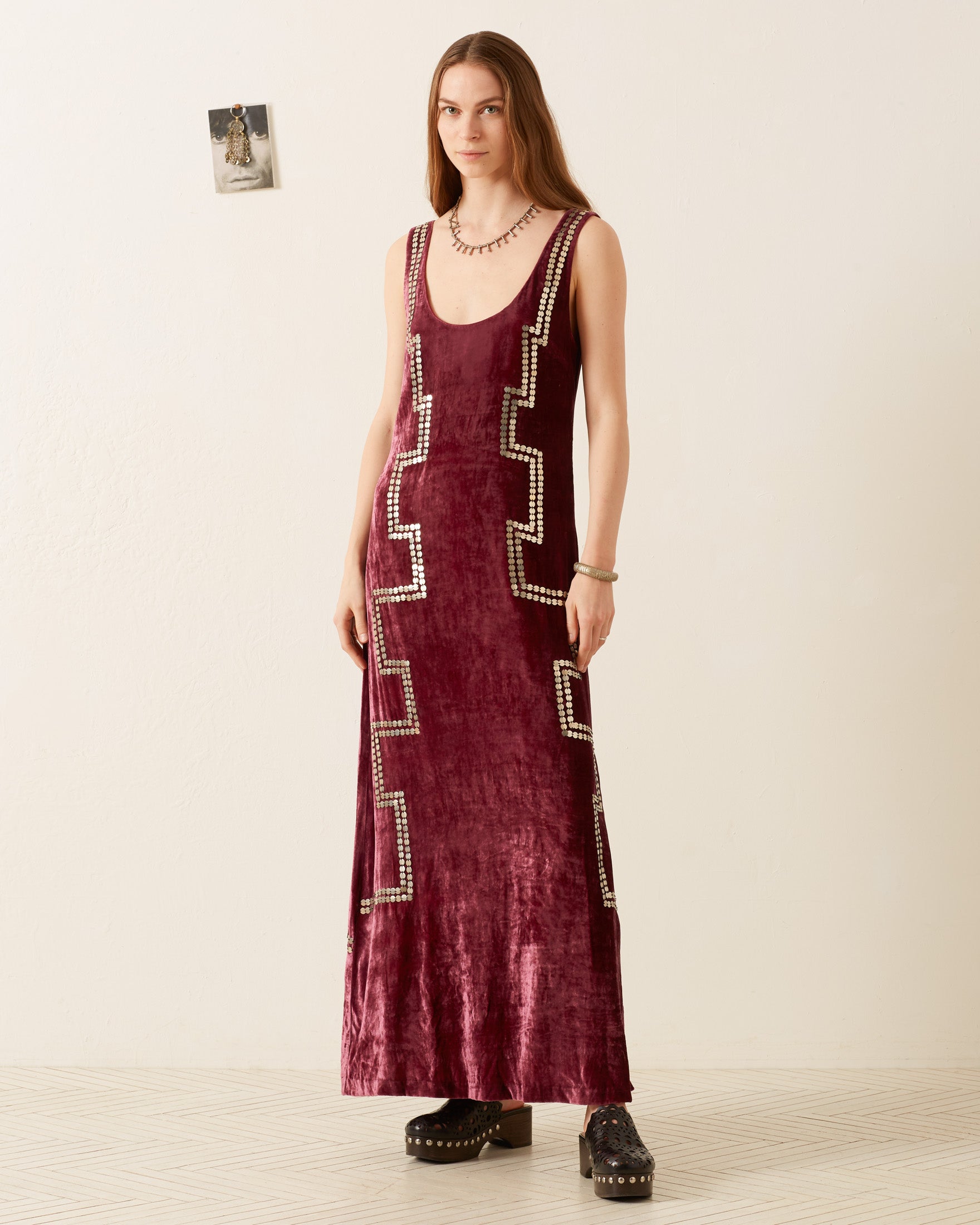 An individual stands against a stark white wall, elegantly dressed in the Adele Plum Velvet Dress, featuring intricate geometric patterns. Their hair flows down as they confidently sport black platform shoes, with a framed picture nearby enhancing the minimalist setting.