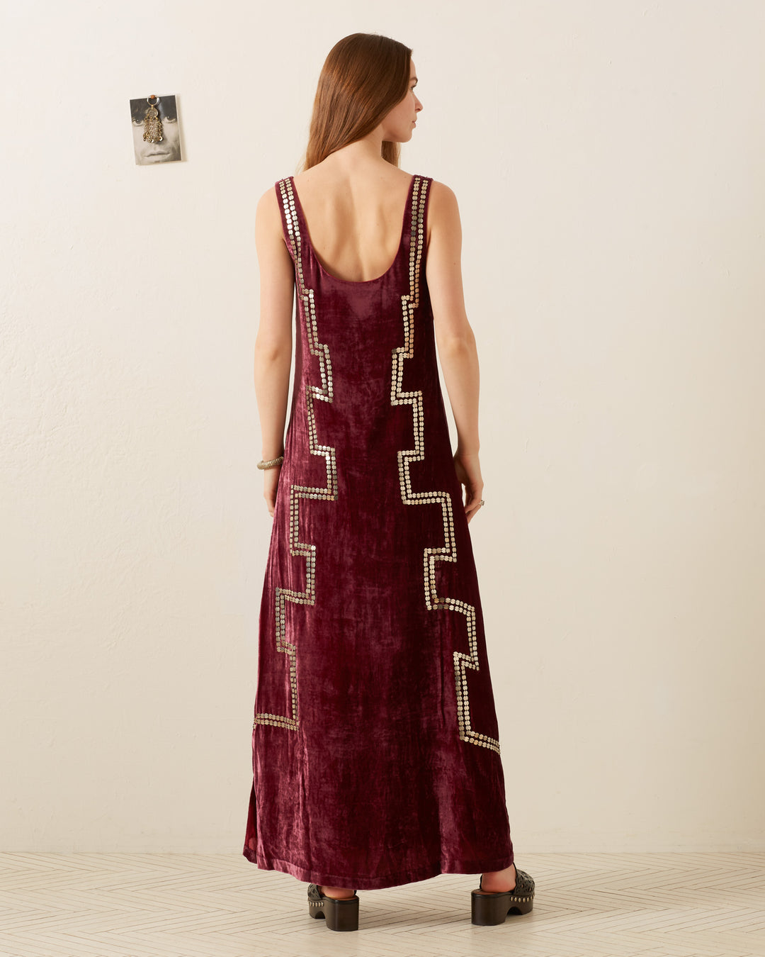 A woman with long brown hair stands with her back to the camera, wearing the Adele Plum Velvet Dress, which is adorned with metallic geometric patterns. She is in a minimalist room with a small photo on the wall.