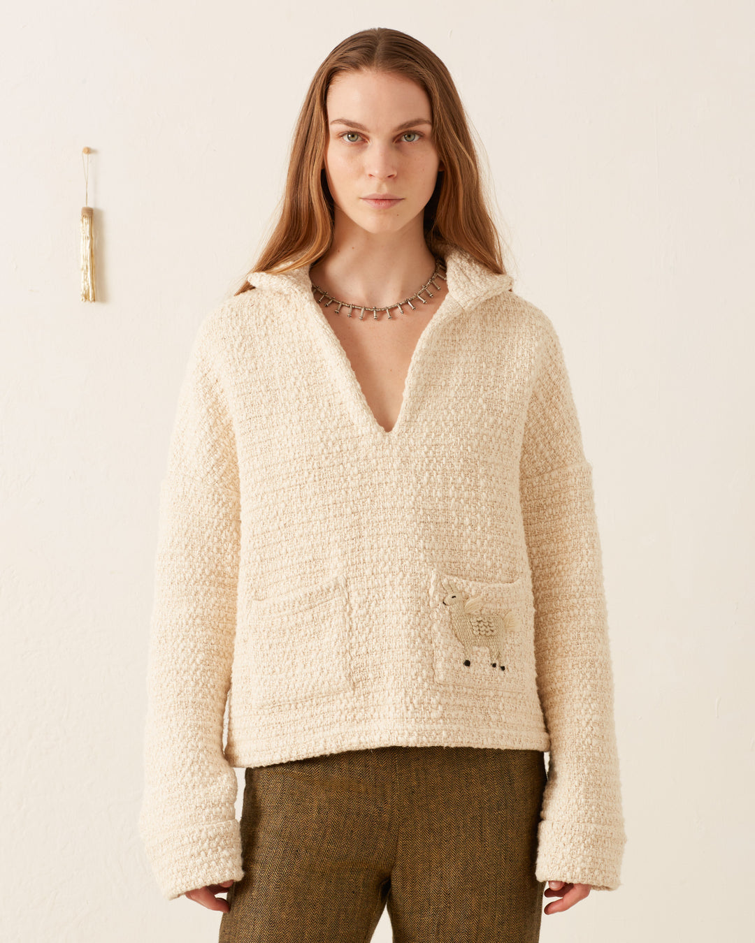 A person wearing the Allegra Pearl Bouclé Top, featuring a cream color with two front pockets and a distinctive handmade llama appliqué, is paired with brown pants and stands against a plain background.