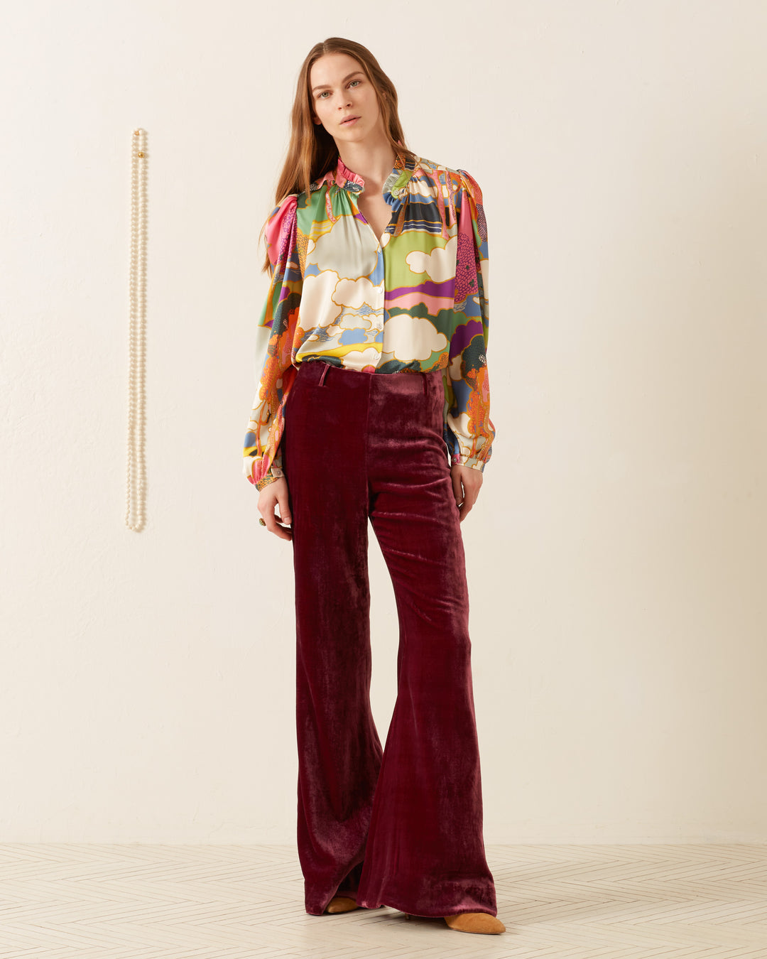 Wearing an Annabel Aurora Silk Shirt, a woman stands against a plain wall, her ensemble completed with red velvet pants and an Italian silk charmeuse scarf elegantly draping beside her.