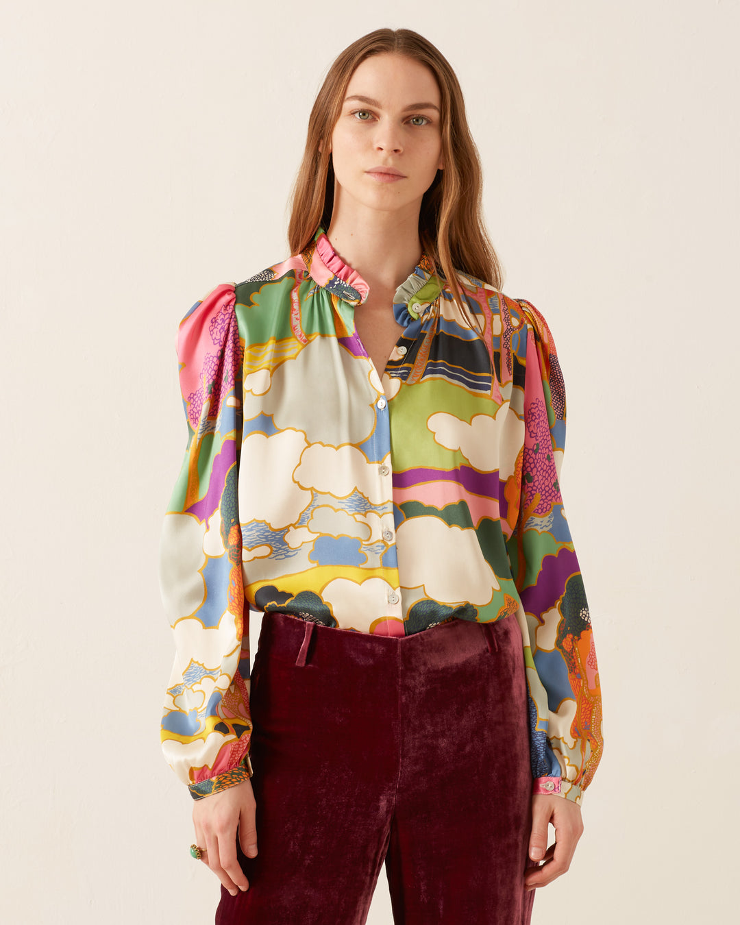 A person wearing an Annabel Aurora Silk Shirt featuring a vibrant Art Nouveau landscape print, paired with velvet pants, stands against a plain background.