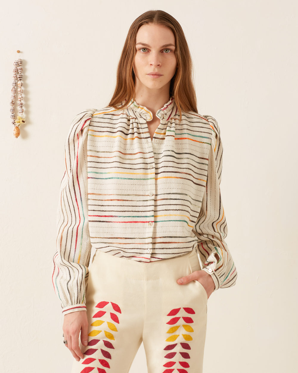 A person wearing an Annabel Chroma Weave Shirt and white pants adorned with colorful leaf patterns stands against a plain background.