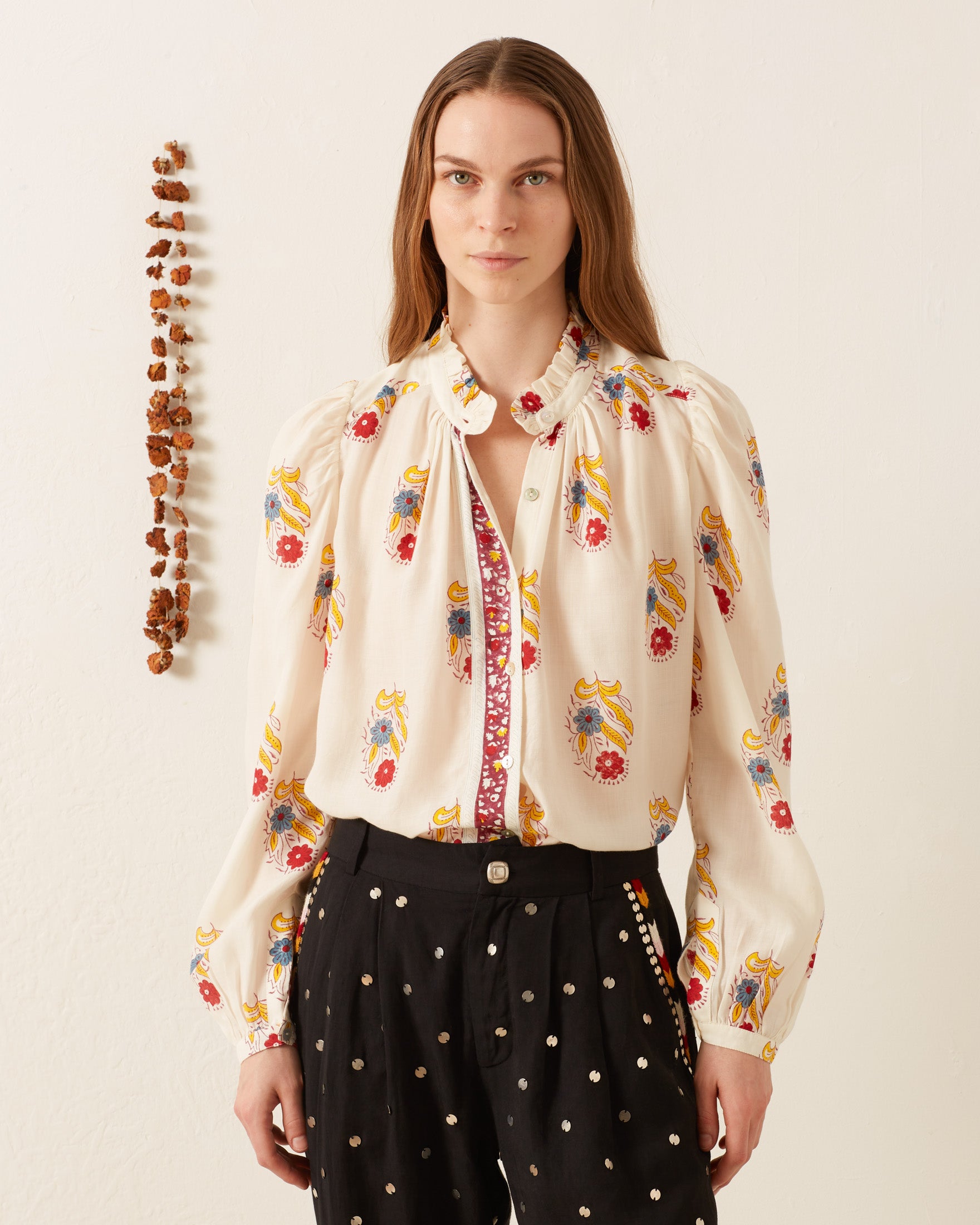 A person wearing an Annabel Fireflower Shirt and polka dot pants stands against a plain background with a vertical line of rocks on the wall.