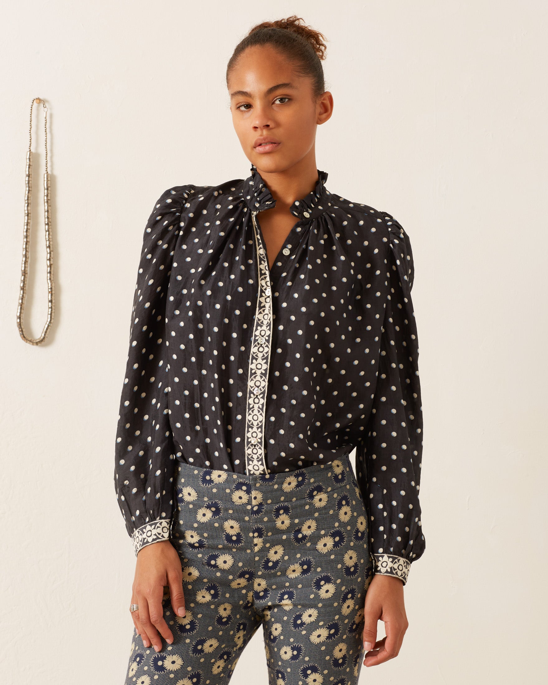 A person stands against a plain light background wearing the Annabel Raven Dot Silk Shirt, featuring intricate hand-block printed polka dots with white decorative trim, along with high-waisted floral patterned pants. A necklace is visible hanging on the wall in the background.