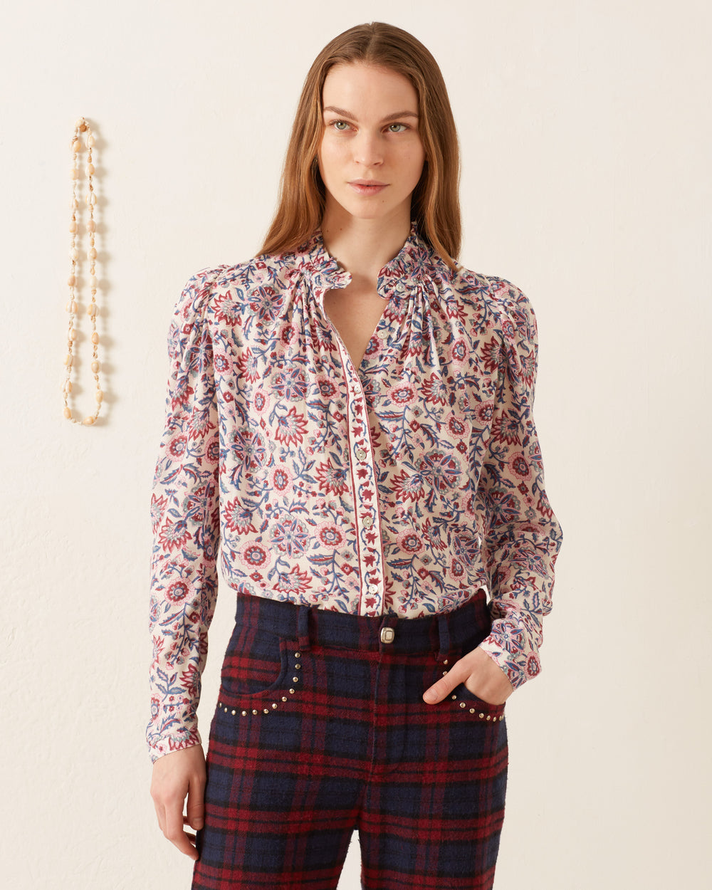 A woman with straight hair stands against a white wall, wearing the Artemis Amazon Floral Shirt, which features a vintage-inspired ruffled collar and high-waisted plaid pants. Her hand rests in her pocket while a beaded necklace hangs beside her.