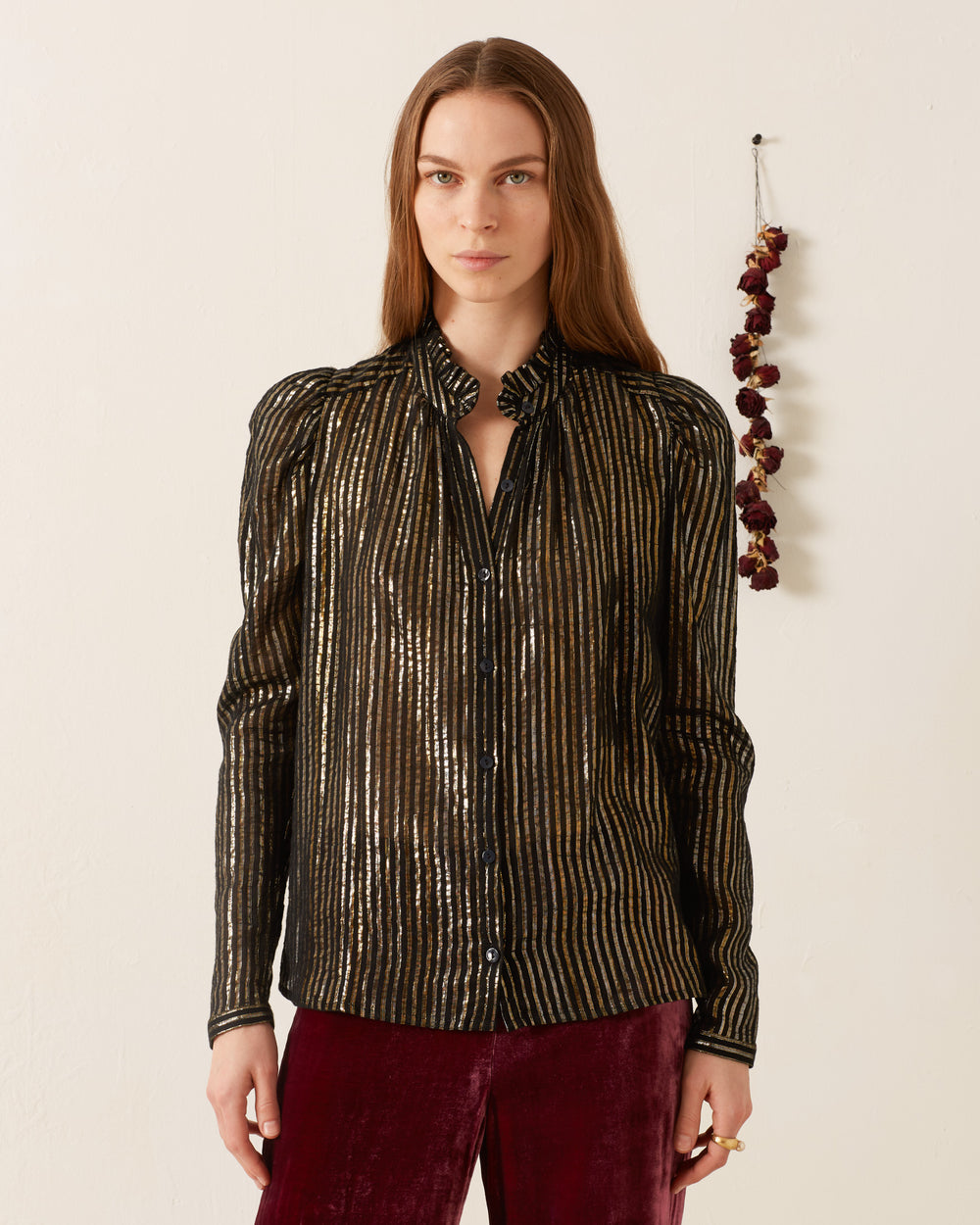 A person with long hair is wearing the Artemis Moonlight Shirt, accented with silver Lurex stripes for a '70s disco flair, paired with maroon pants. They stand in a minimalist setting, where a small bunch of dried flowers hangs on the wall behind them.