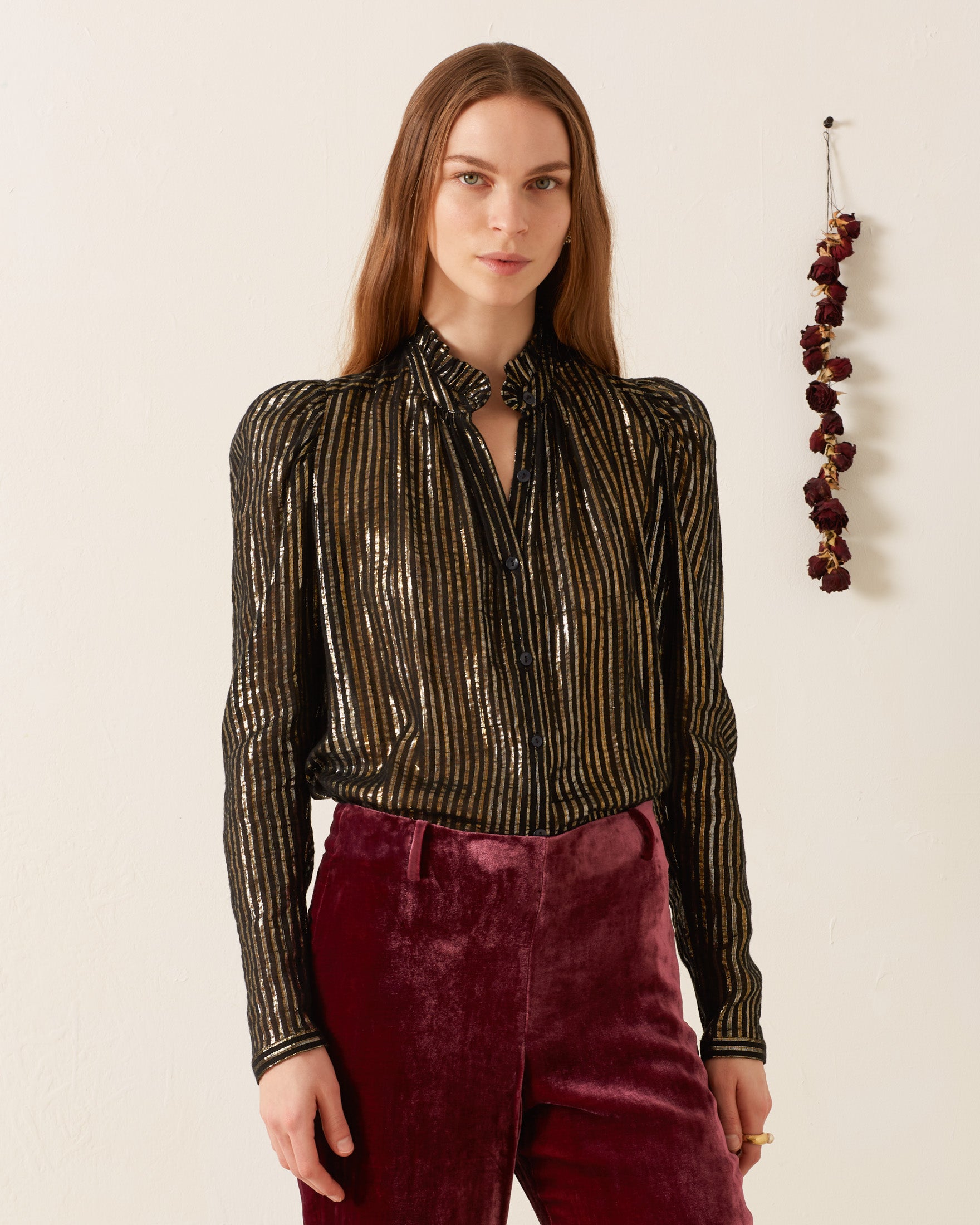 A person stands against a white wall, exuding '70s disco flair in the Artemis Moonlight Shirt paired with purple velvet pants. Dried flowers hang on the wall behind them, adding an artistic touch to the scene.