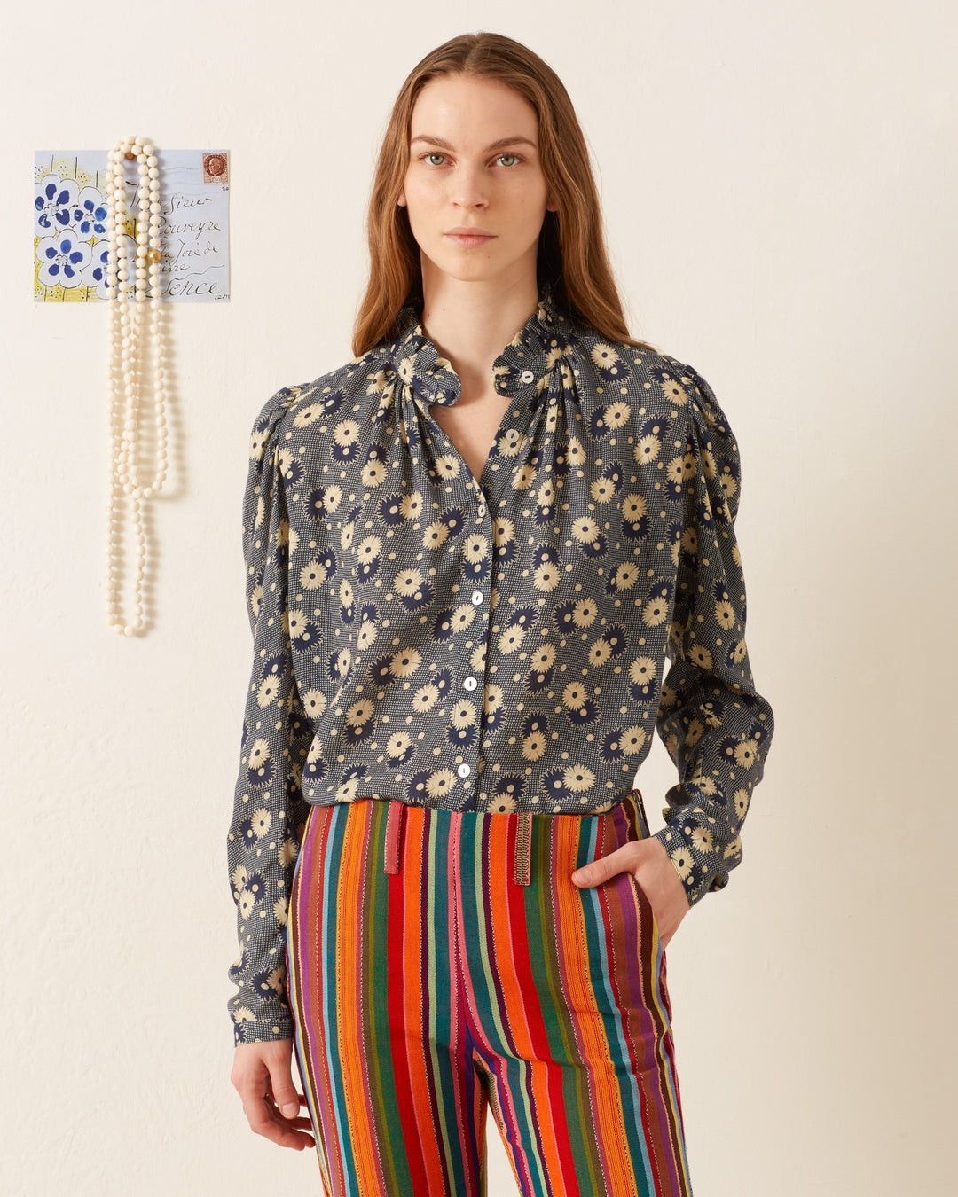 A person with long hair wearing an Artemis Navy Daisy Shirt and colorful striped pants stands against a plain backdrop, with a string of pearls and a small piece of illustrated paper pinned to the wall in the background.