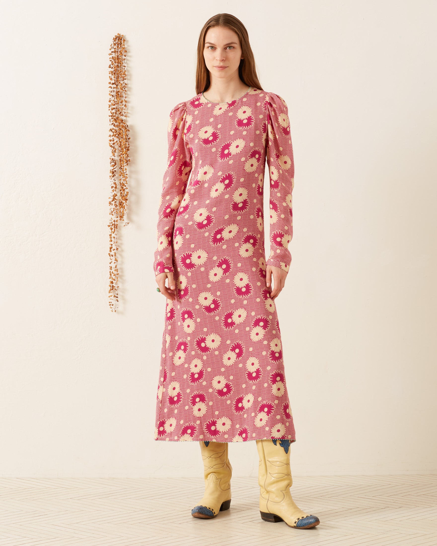 A person stands wearing the Astrid Berry Daisy Dress, a vintage-inspired pink floral dress, paired with yellow cowboy boots against a white textured background. A vertical decorative element hangs on the wall, creating a minimalist and stylish setting that exudes a '70s-style vibe.