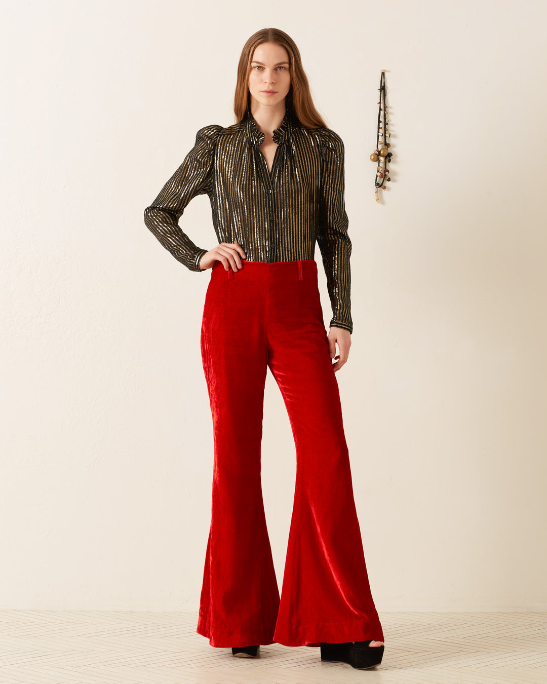 A woman stands confidently in a black and gold striped blouse, paired with the 'Charlie Cherry Velvet Pant' featuring a '70s-style red velvet flare. Her long brown hair flows elegantly as she poses against a simple white wall adorned with a decorative hanging in the background, exuding retro charm.