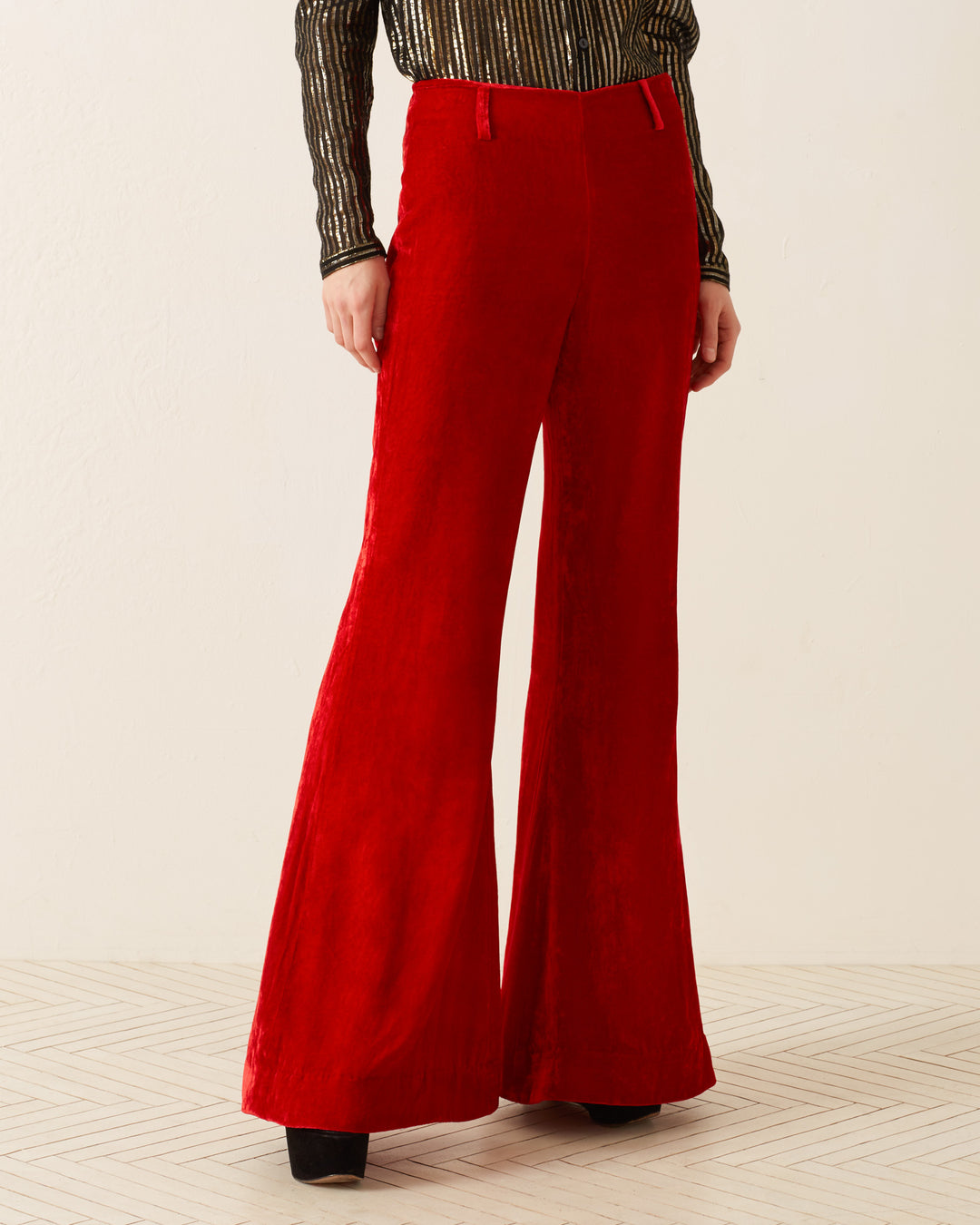 A person is wearing Charlie Cherry Velvet Pants, designed with a '70s-style featuring luxurious red velvet fabric. These high-waisted pants with wide flared legs are paired with a black striped long-sleeve shirt, making a bold statement against the light-colored wall. The subtle herringbone pattern on the floor adds an elegant touch to the scene.