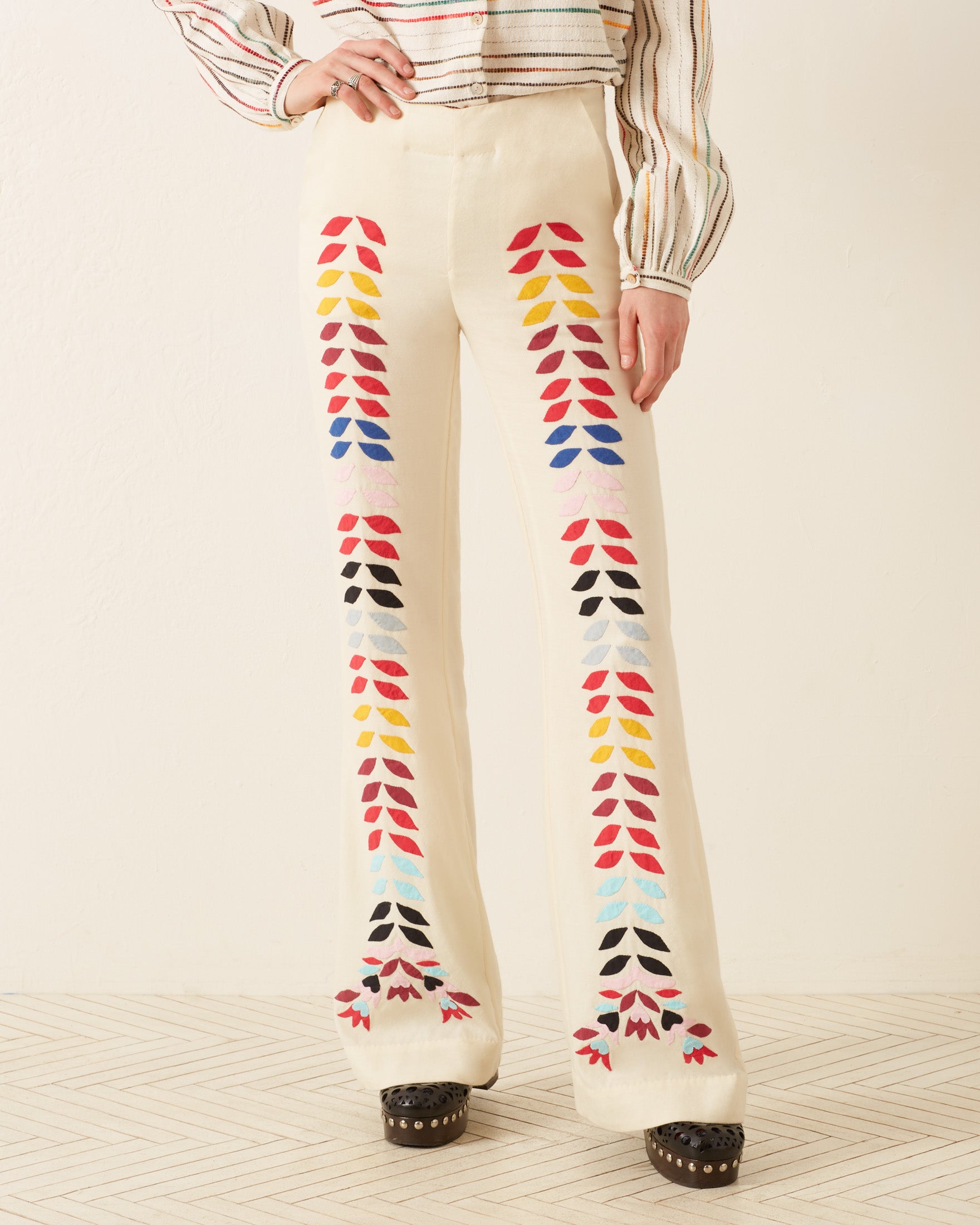 A person wearing the Charlie Ivory Prism Pant, featuring a '70s-style design with colorful leaf patterns down the front, stands on a light geometric floor. The pants are paired with a long-sleeve striped top and black platform shoes, adding a touch of retro flair to the ensemble.