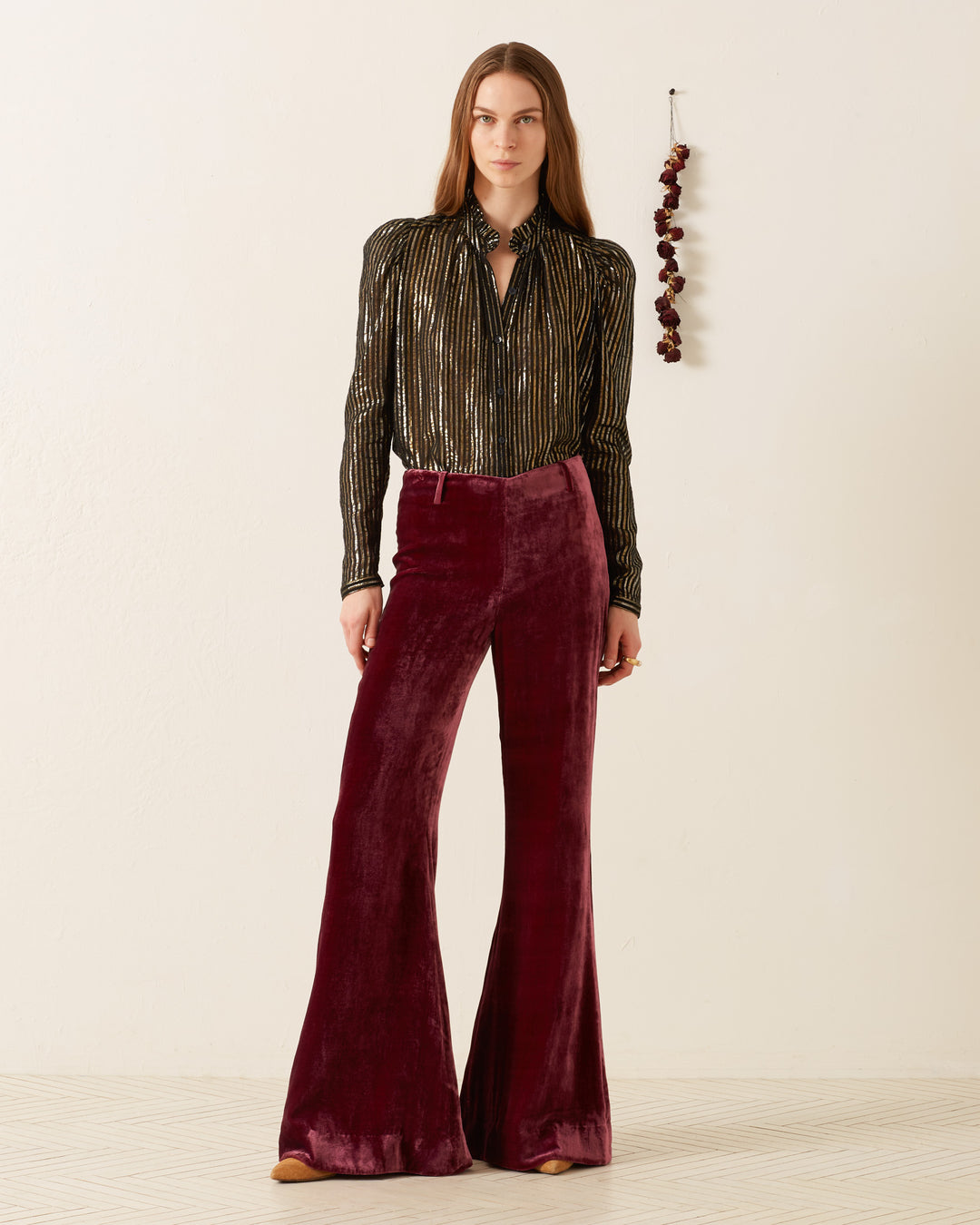 A person stands against a plain wall, embracing '70s style with a shiny, striped blouse and wearing the Charlie Plum Velvet Pant featuring maroon bell-bottom flare legs. To their right, a cluster of small, hanging dark-red objects adds an intriguing touch.