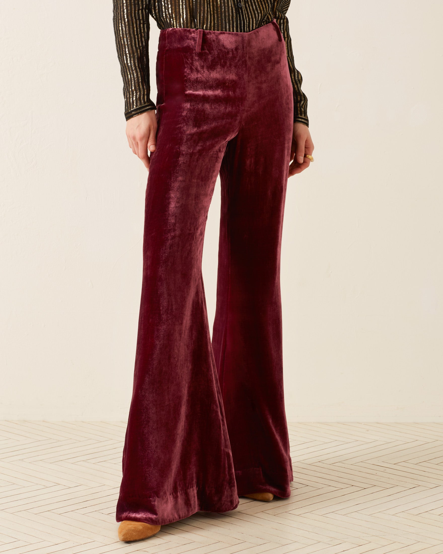 A person is embracing '70s style, wearing high-waisted, Charlie Plum Velvet Pant with bell-bottom flare and a long-sleeved top with a black and gold metallic striped pattern. The beige herringbone floor contrasts with the light-colored wall in the background.