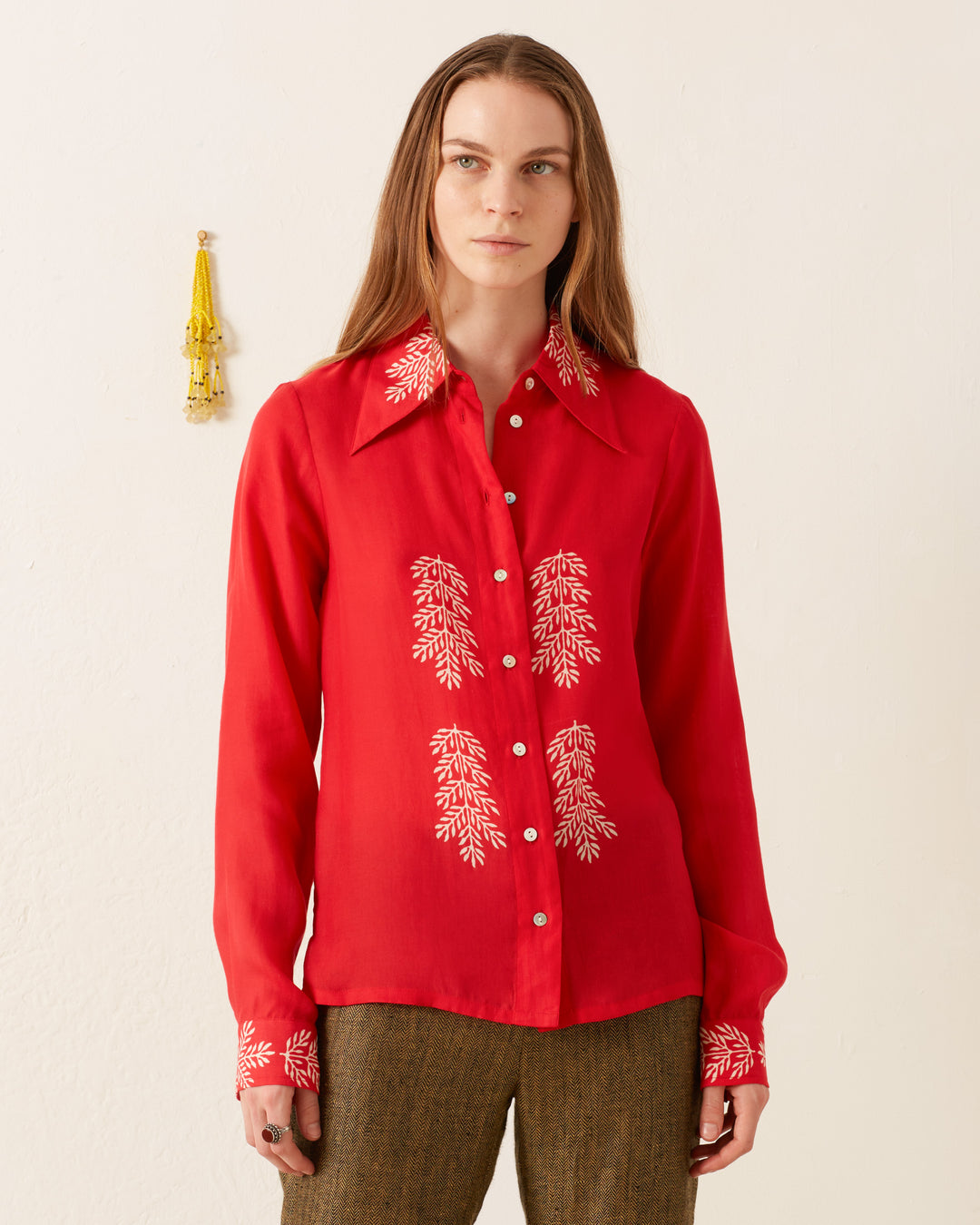 A person with long hair wears the Dolly Cherry Mint Shirt, a vintage-inspired red button-up featuring white floral embroidery on the collar, chest, and cuffs. They stand against a plain white wall adorned with a small yellow decorative piece.