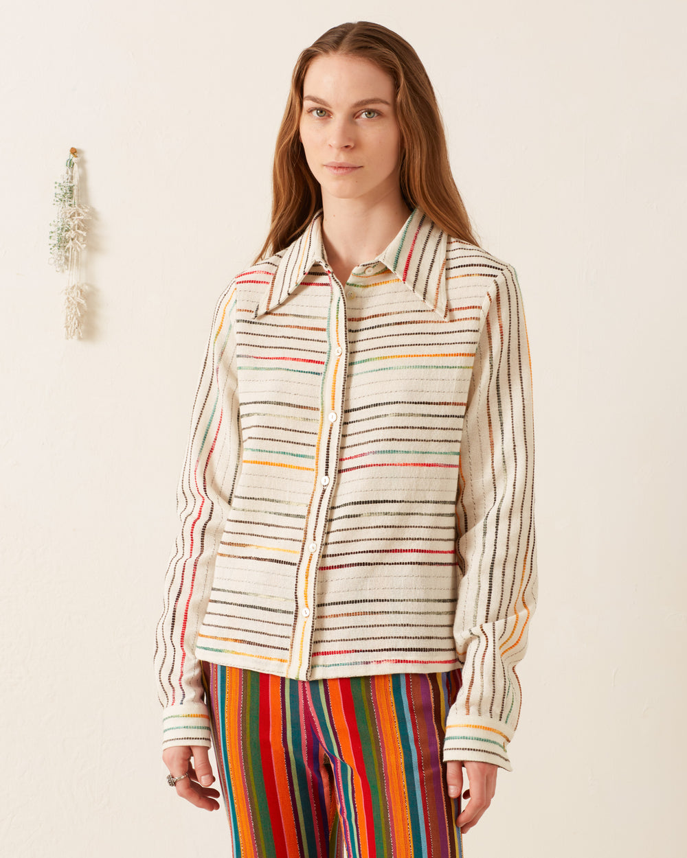 A person wearing the Dolly Chroma Weave Shirt, characterized by its vintage-inspired multicolored stripes and '70s-style wing collar, along with matching pants, stands against a plain wall, complemented by a small decorative hanging plant.