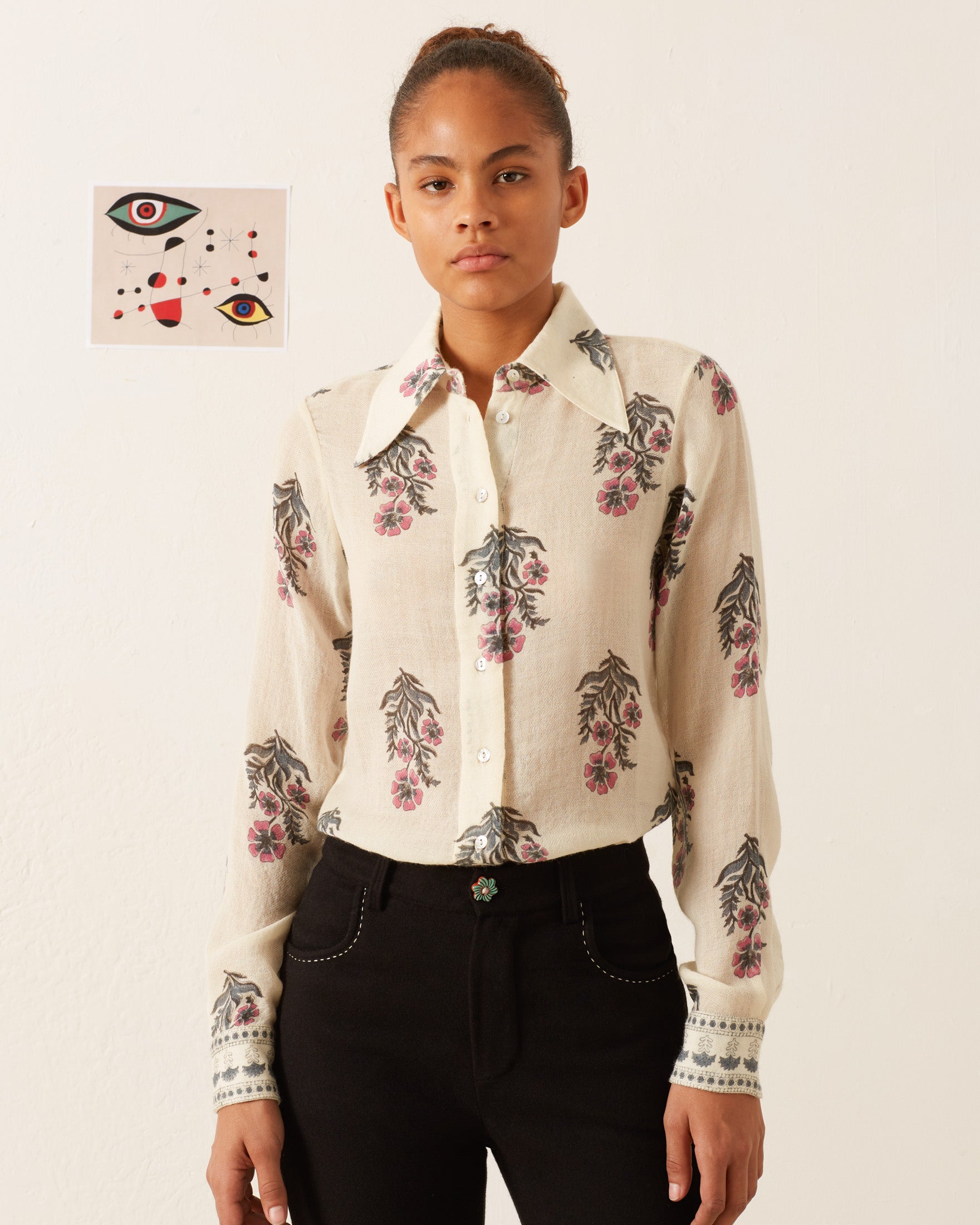 A person with a neutral expression stands against a plain background, wearing the Dolly Mirabilis Wool Shirt with a long-sleeve, vintage-inspired floral motif and black pants. An abstract artwork featuring eyes and geometric shapes is hung on the wall behind them.