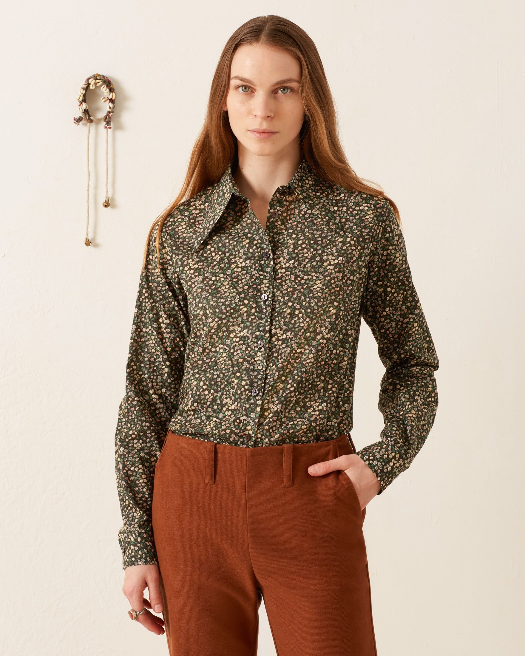 A woman stands against a plain background wearing the vintage-inspired Dolly Moonflower Shirt and brown pants, with her hand in her pocket. A decorative object is on the wall.