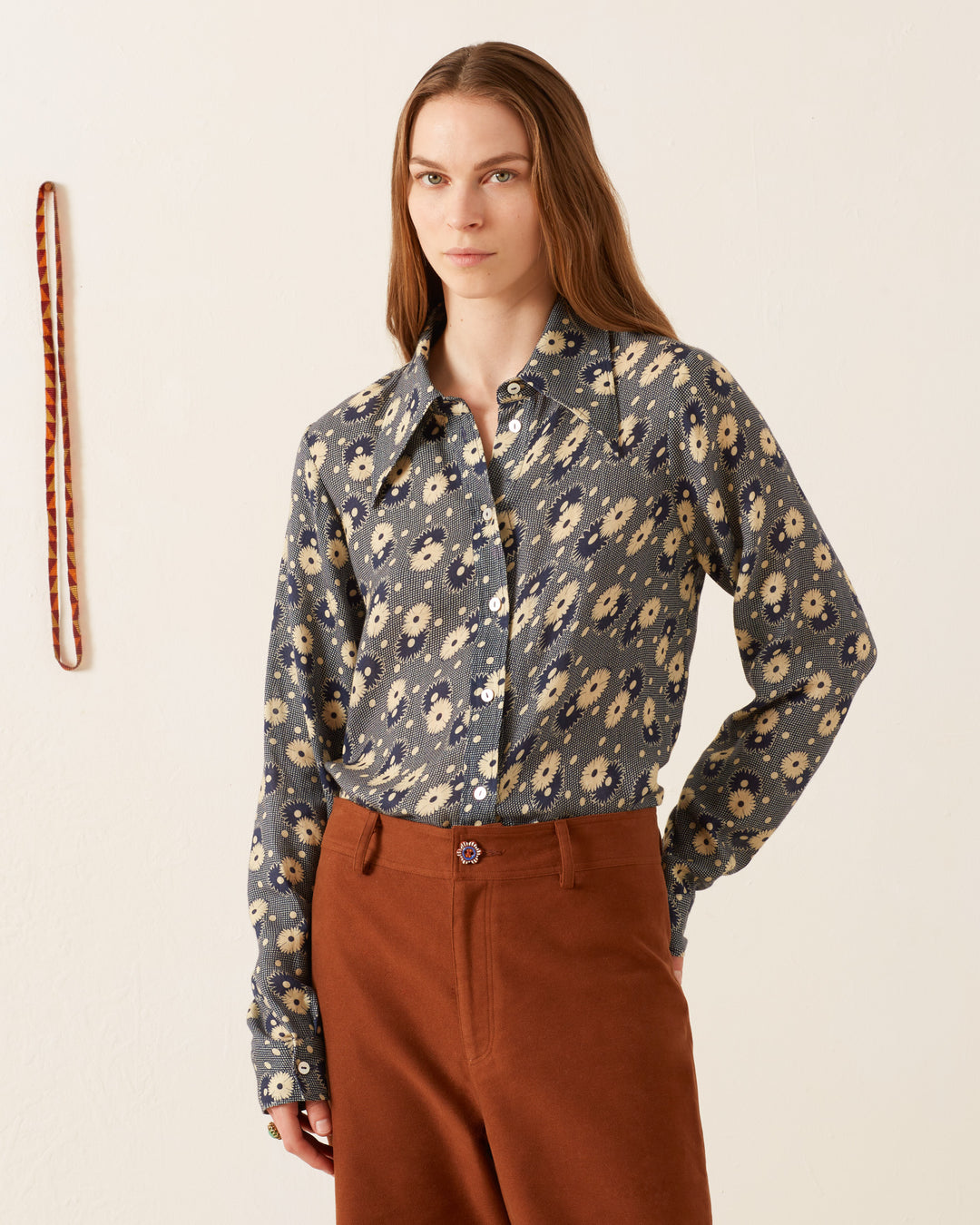 A person with long hair wears a '70s-style Dolly Navy Daisy Shirt and brown pants, standing against a plain wall with a red-striped rope hanging.