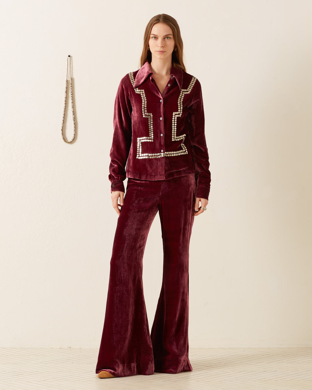 A person dressed in the Dolly Plum Velvet Shirt with bell-bottom pants and a vintage-inspired, hand-embellished style stands in a minimalist room where a necklace is displayed on the wall.