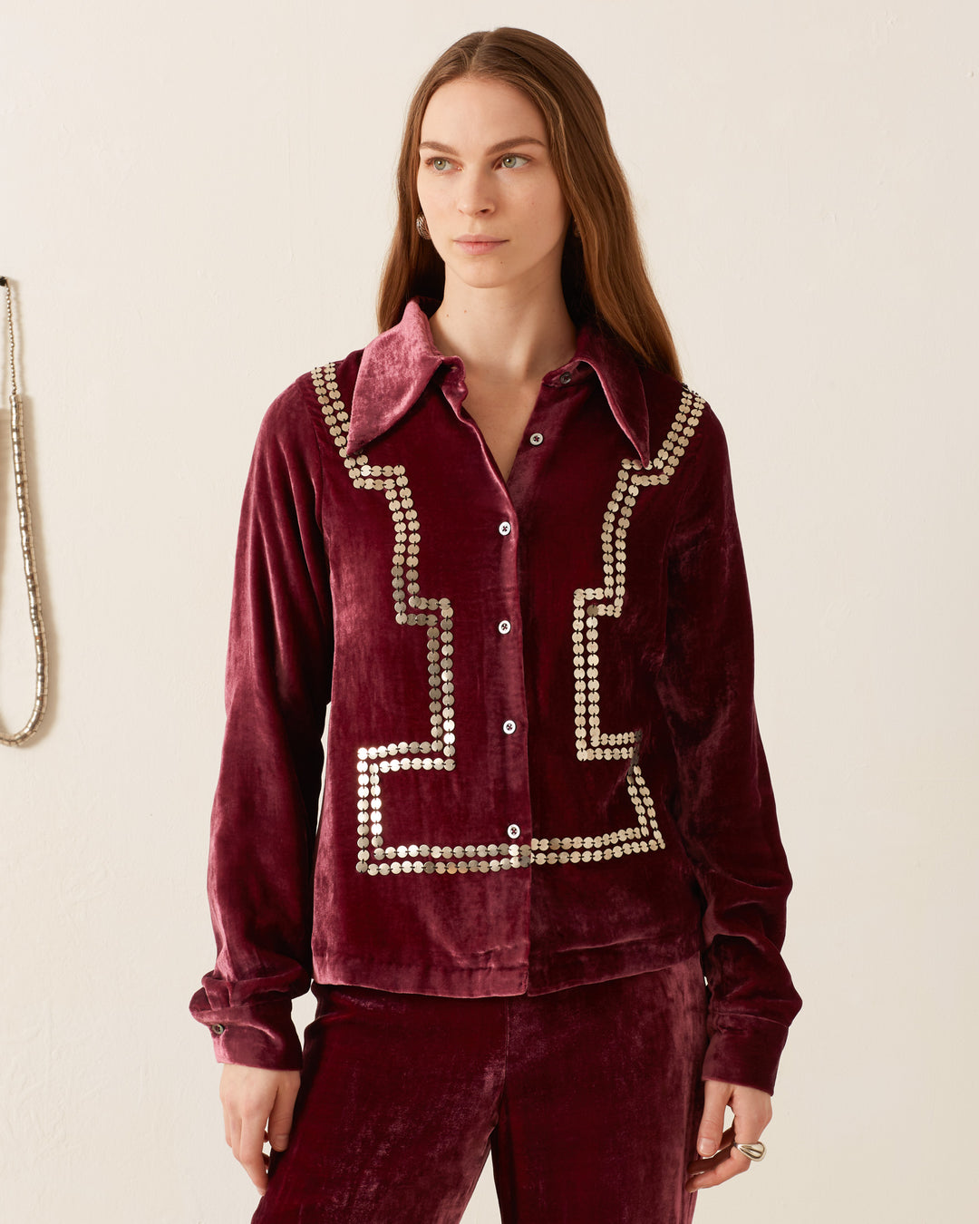 Against a light background, a woman stands wearing the Dolly Plum Velvet Shirt, which features a vintage-inspired design in burgundy velvet with white decorative patterns.