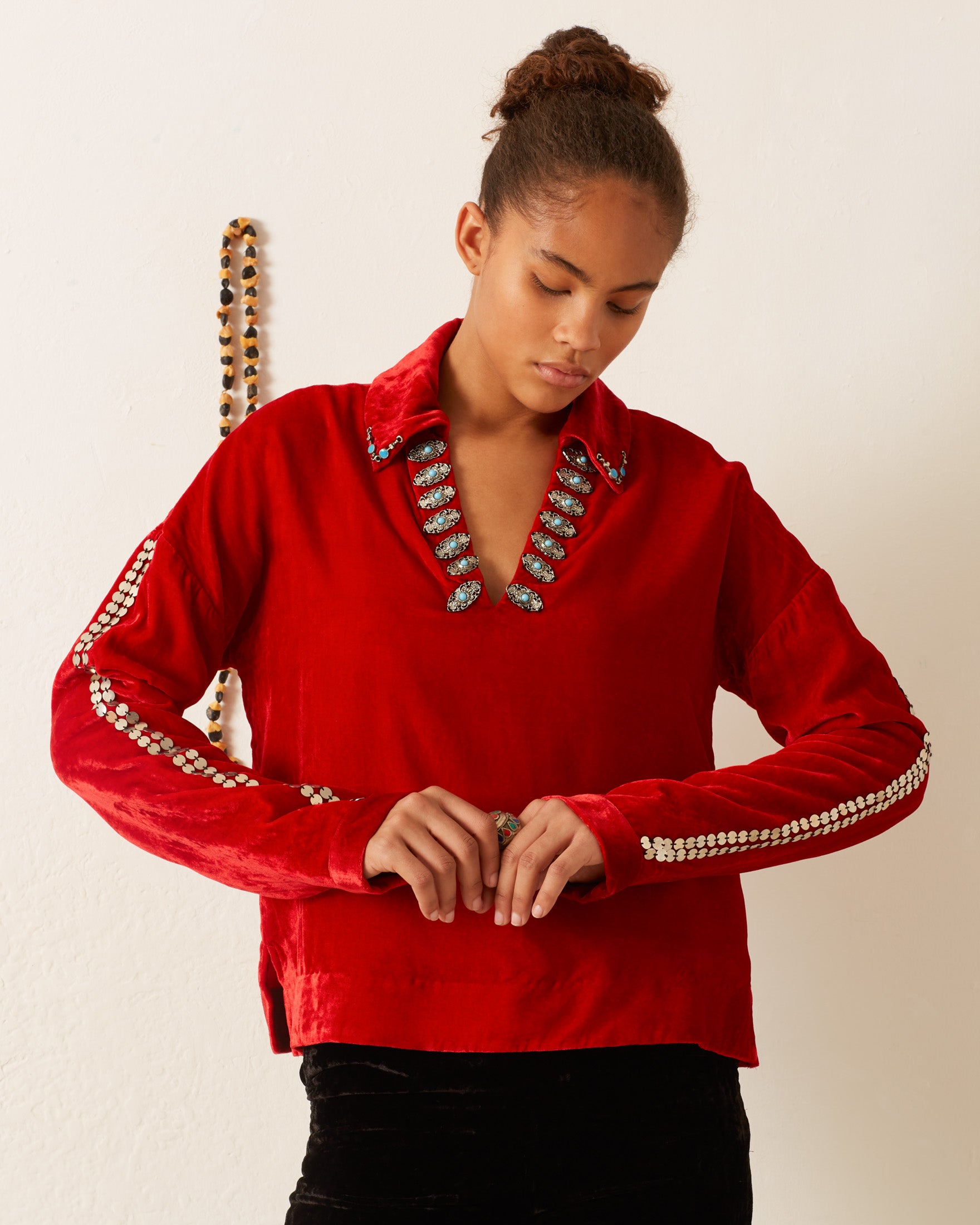 A person wearing the Dylan Ruby Velvet Tunic, adorned with decorative beading on the collar and sleeves, stands near a white wall with hanging beads.