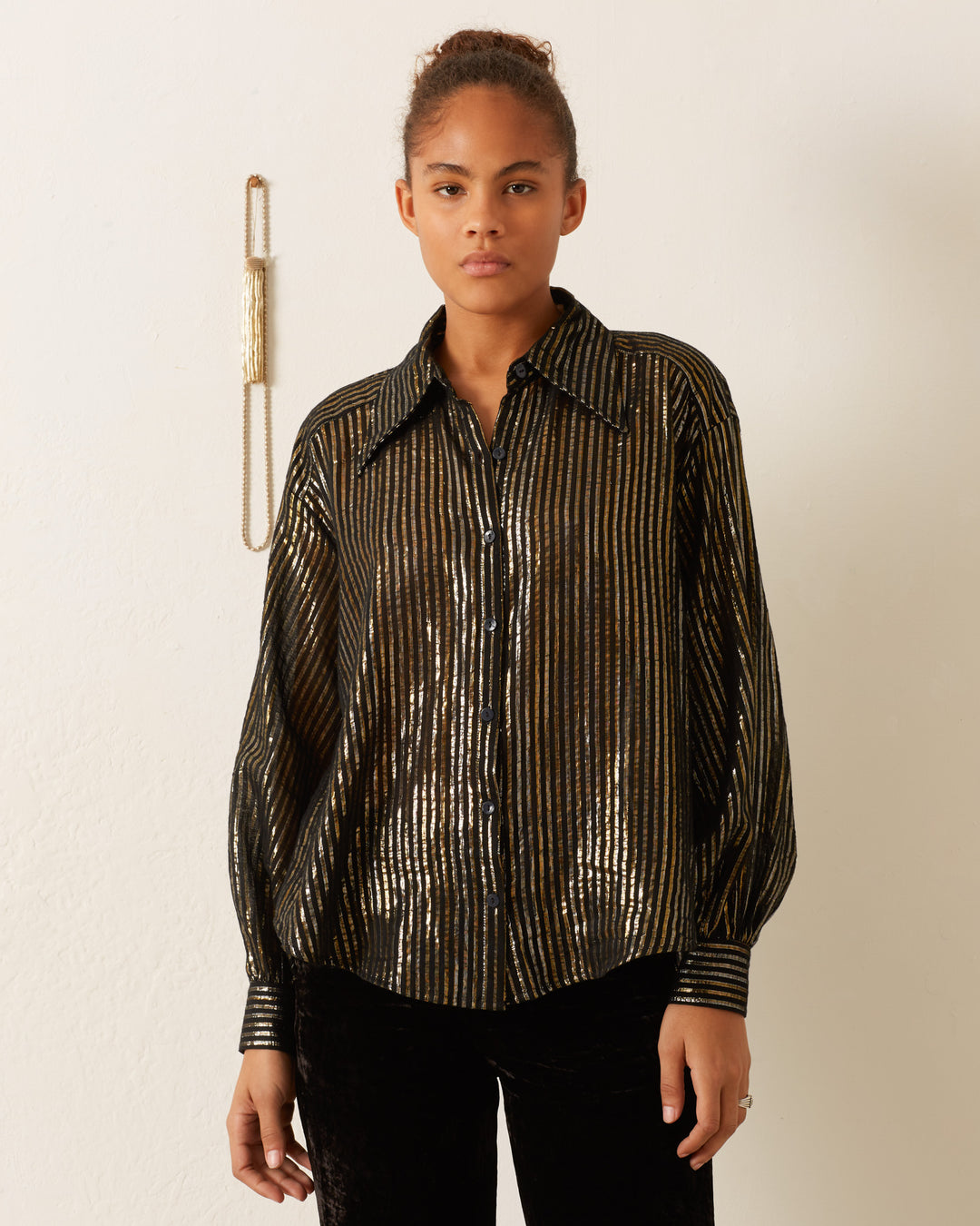 Dressed in an Emery Moonlight Shirt, featuring a shimmering black and gold metallic stripe with a '70s-style wing collar, an individual stands against a light-colored wall.