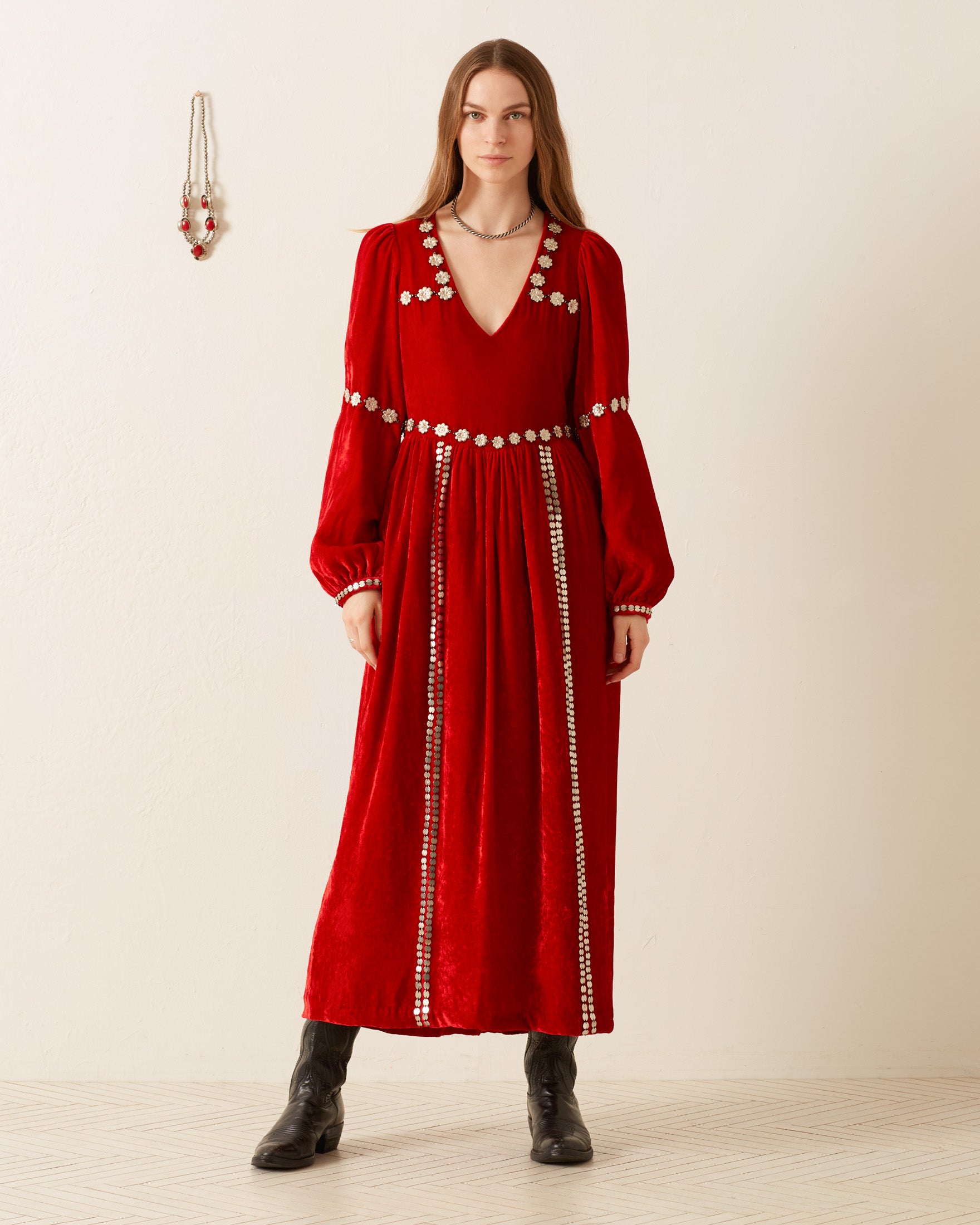 A woman stands against a plain background, wearing the Imogen Ruby Velvet Dress, adorned with floral embroidery along the neckline and waist. She pairs this stunning dress with black boots. A necklace is displayed on the nearby wall.