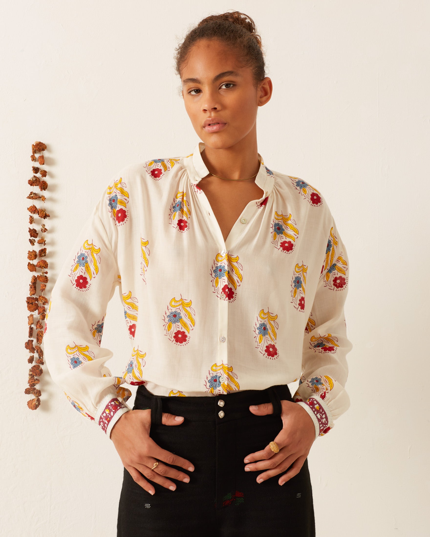 A person wearing the vibrant Kiki Fireflower Shirt, featuring colorful floral motifs, and black pants stands against a plain background. A vertical string of small, brown objects hangs on the wall to the left, adding texture to the scene.