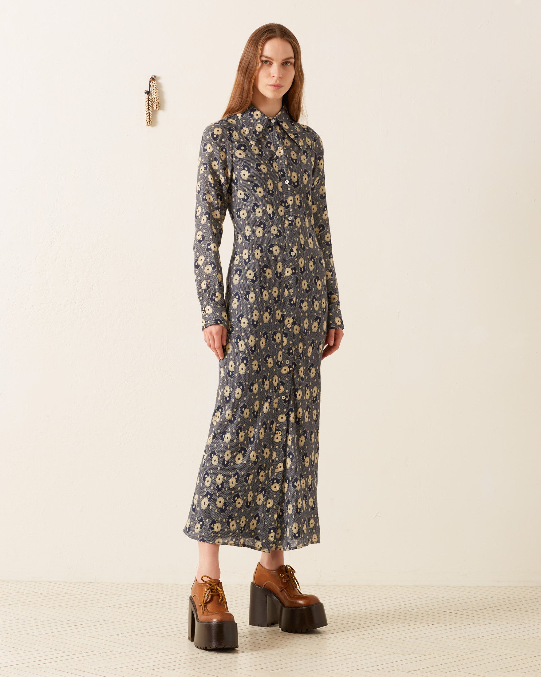 A person stands against a plain wall wearing a Lotte Navy Daisy Dress featuring a '70s-style wing collar. They have long hair and are wearing brown platform shoes, adding to the vintage feel. A decorative wall piece is visible in the background.