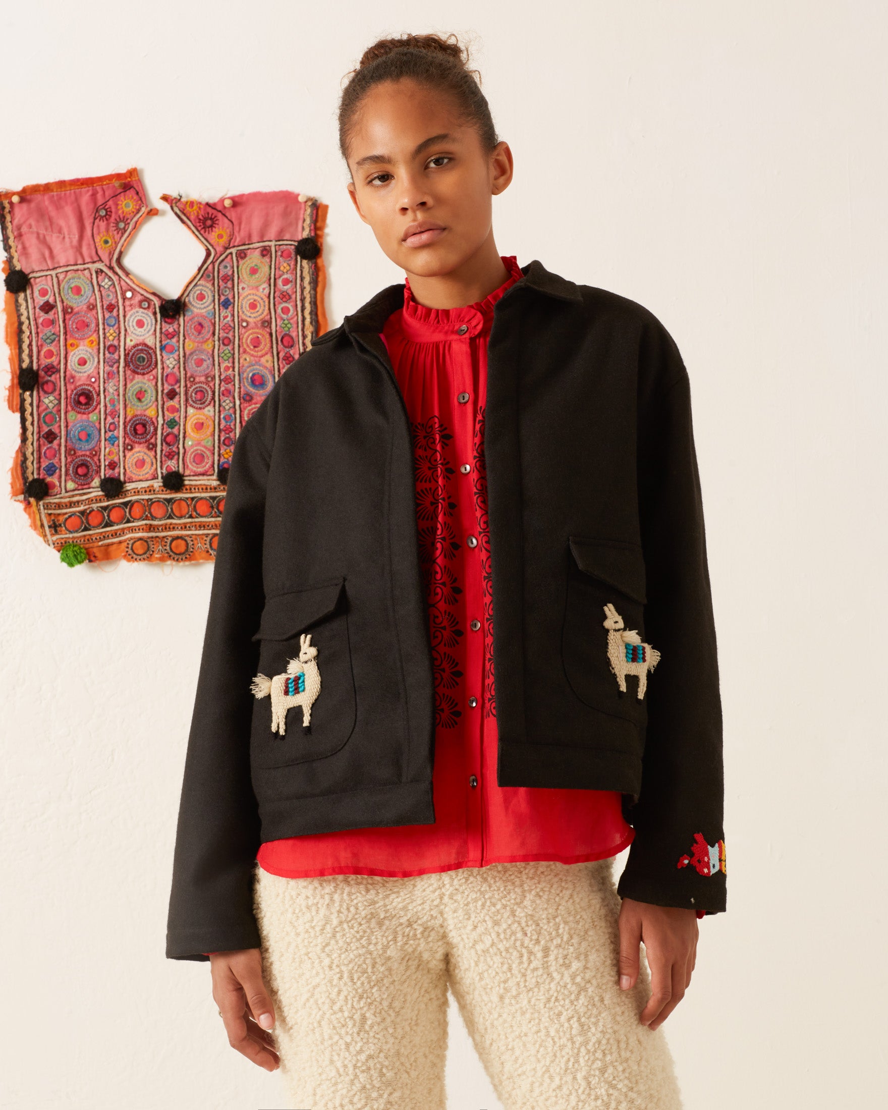 A person wearing a Lucky Little Llama Jacket, paired with a red blouse and woolly pants, stands before a colorful textile hanging on the wall.