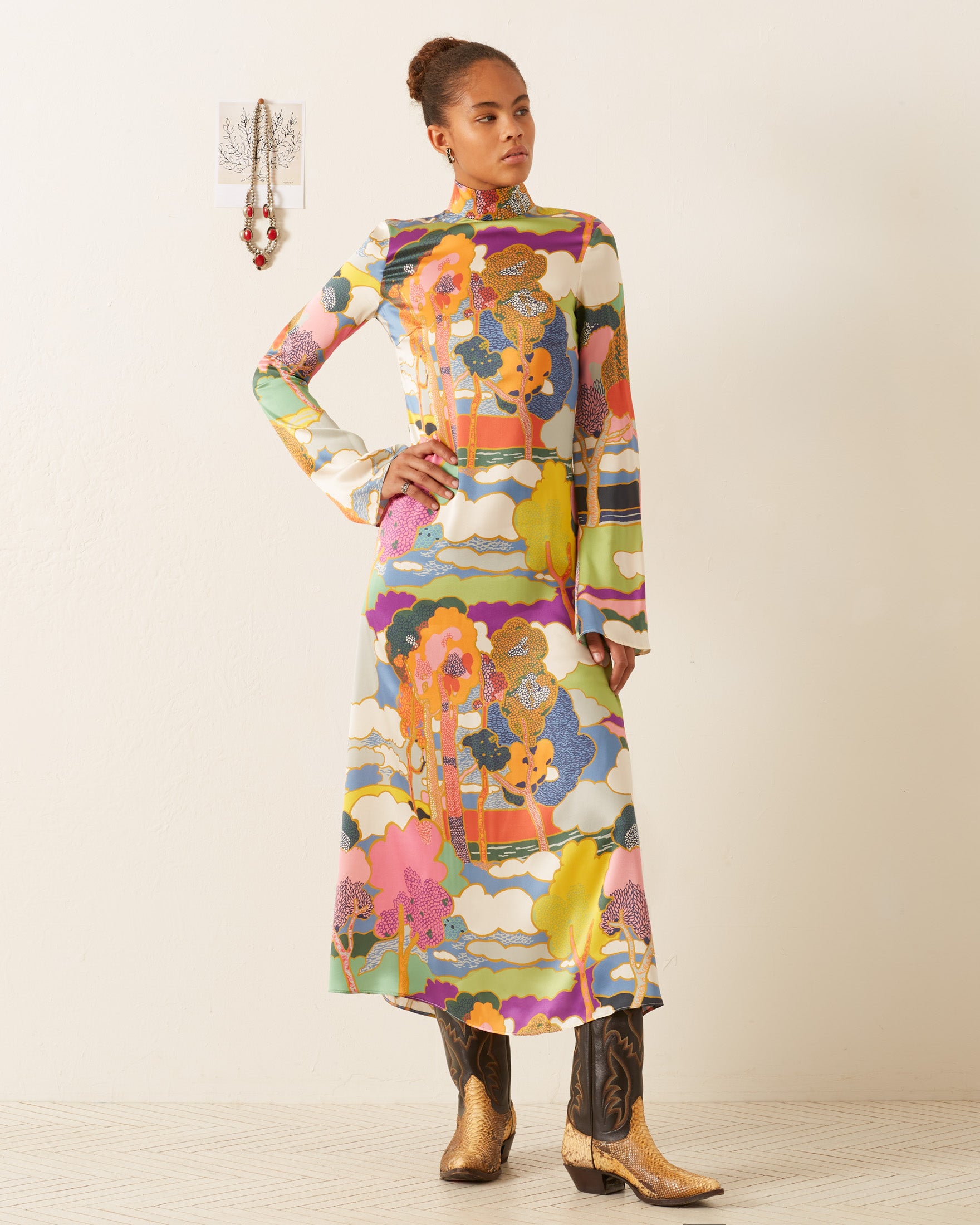 A person stands confidently in the vibrant Nia Aurora Silk Dress, paired with brown cowboy boots. Their hair is styled in a bun, set against an off-white wall adorned with a small decorative item.