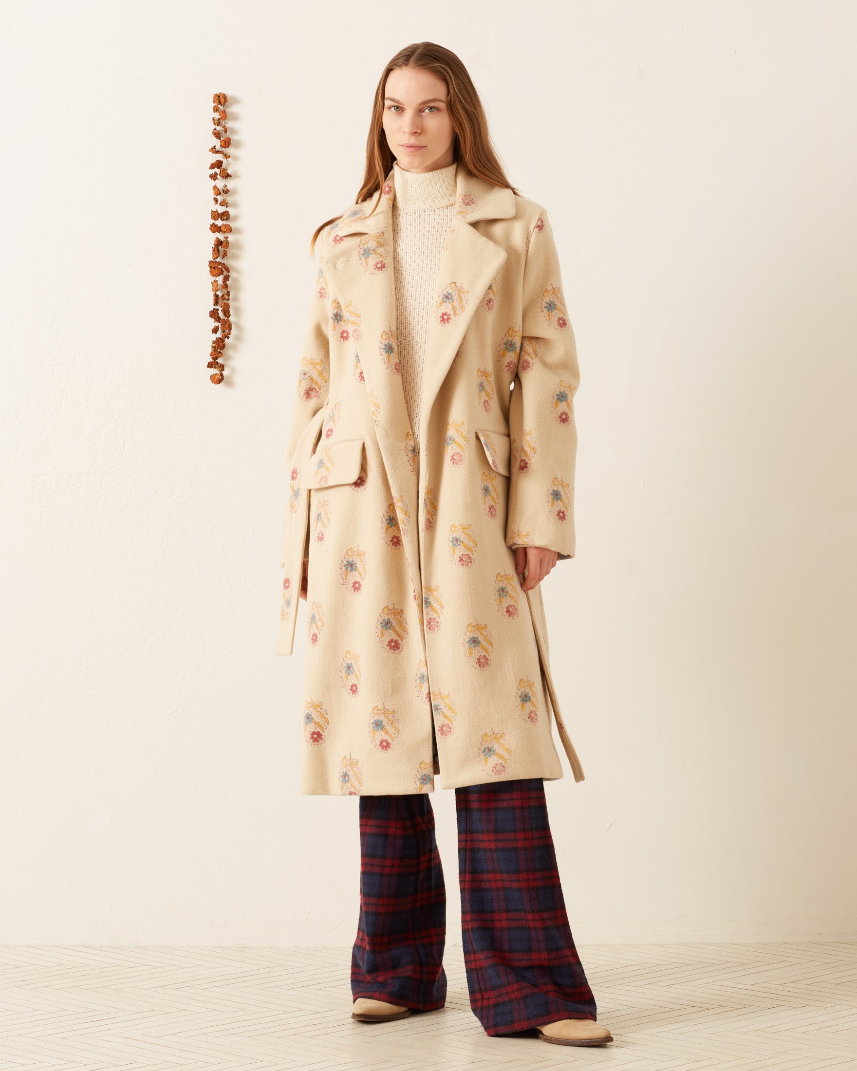 A person stands against a minimalist cream-colored wall, wearing the Officer Fireflower Wool Coat in hand-block printed light beige wool over a white turtleneck and plaid pants. The outfit is complemented by beige shoes, and there is a vertical row of stones on the wall beside them.