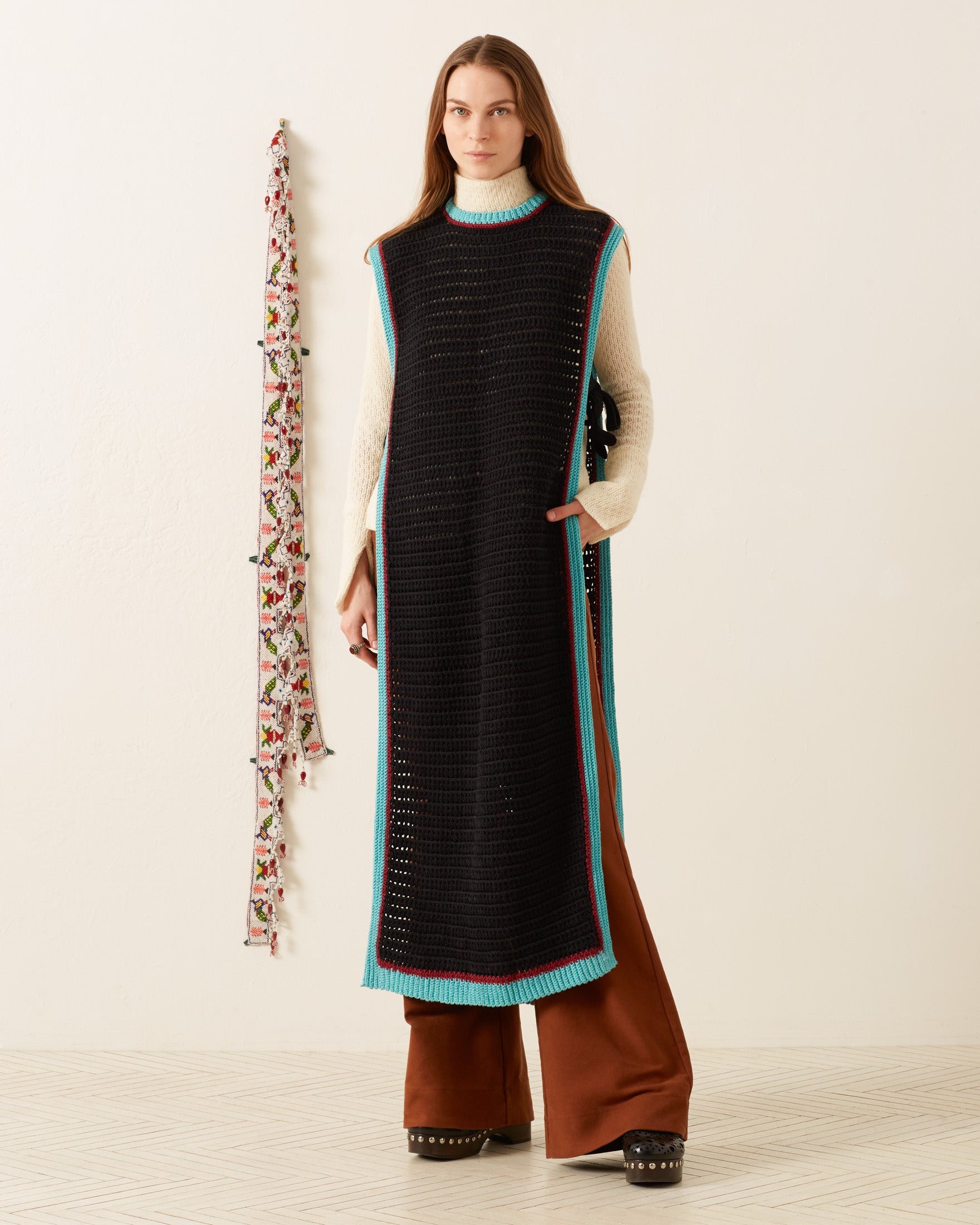 A woman stands indoors in the Omer Equinox Crochet Top—a long, sleeveless black tunic with blue trim—over a cream turtleneck and rust wide-leg pants, beside a floral wall hanging.