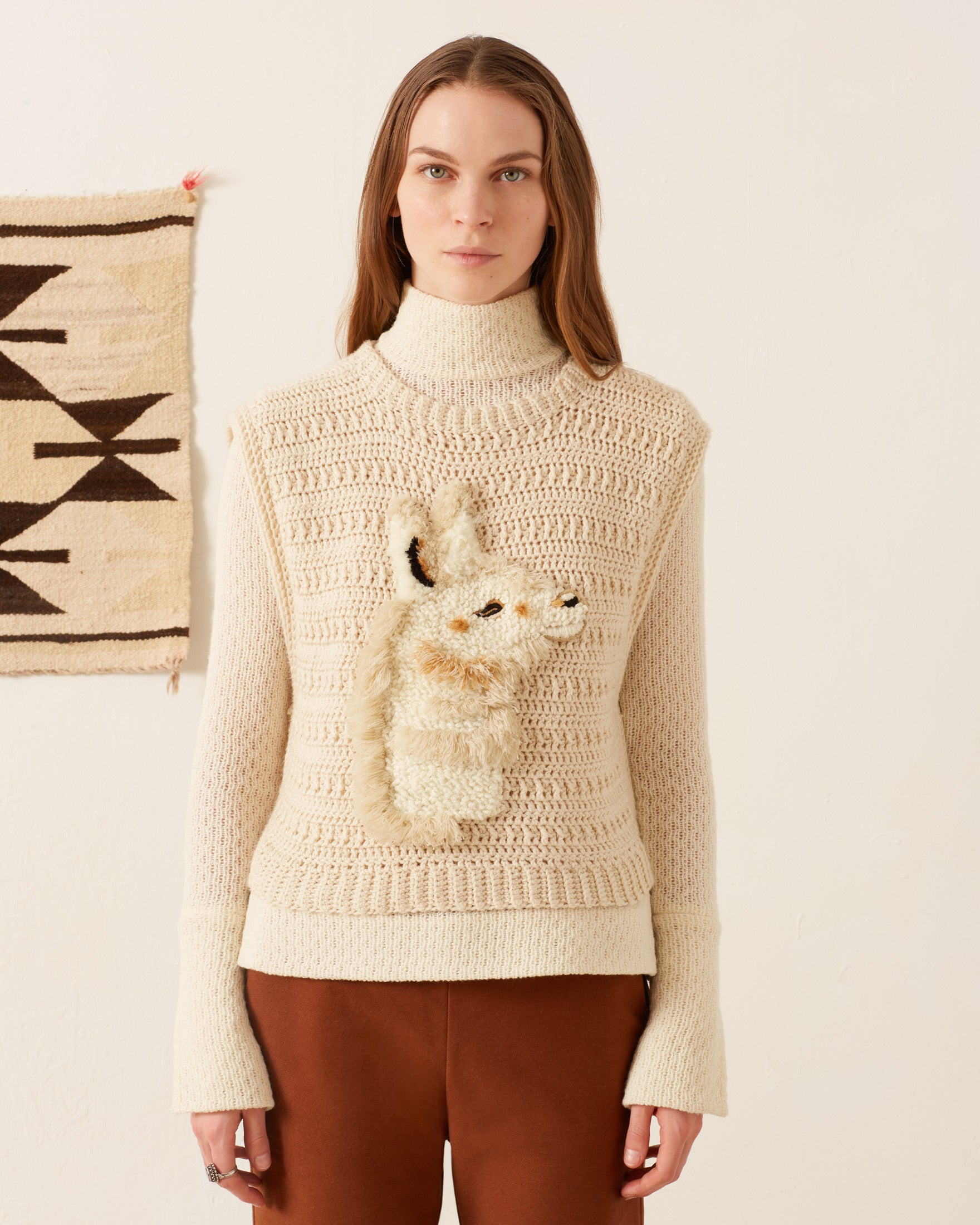 A person with long hair wearing the Oona Llama Crochet Top, which showcases a textured animal design on the front in a cream knit style, stands against a plain backdrop along with a geometric-patterned wall hanging. They complement their vintage-inspired look with brown clothing for the lower half of their outfit.