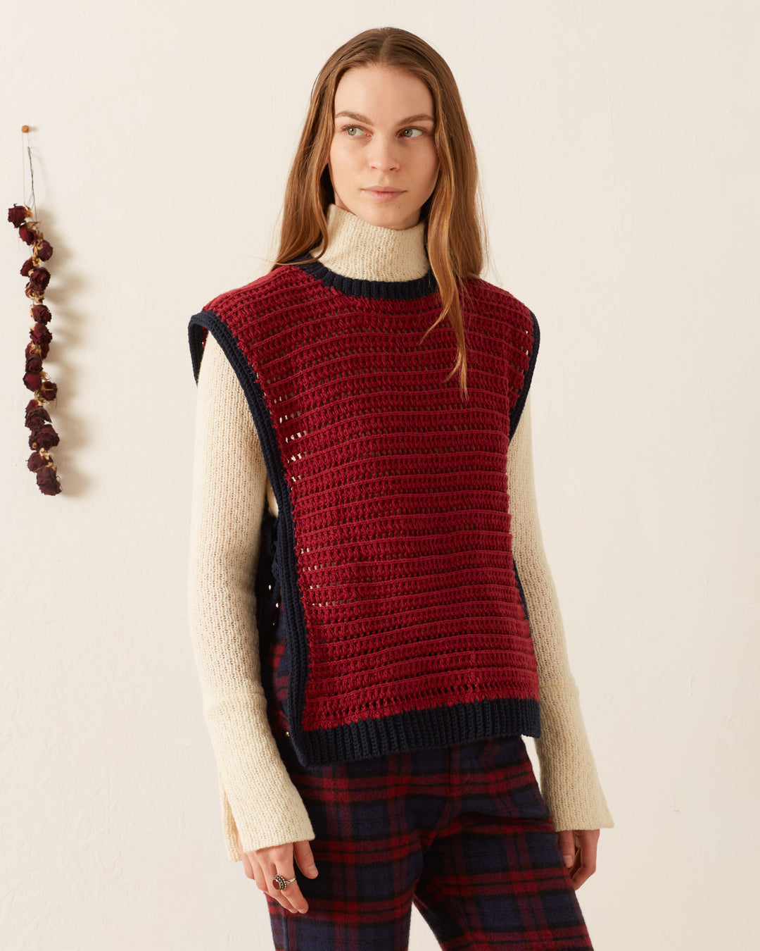 A woman wears the Oona Mulberry Crochet Top over a cream turtleneck sweater with plaid pants, standing against a plain light wall accented with dried flowers.