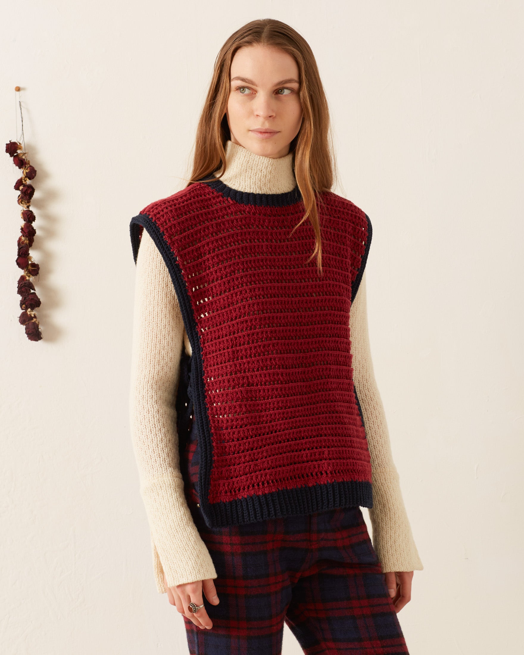 A woman wears the Oona Mulberry Crochet Top over a cream turtleneck sweater with plaid pants, standing against a plain light wall accented with dried flowers.
