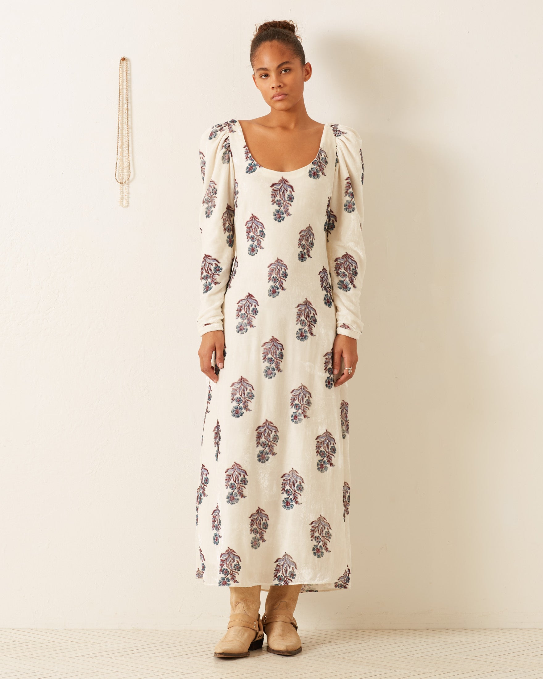 A person stands against a white wall wearing the Paloma Azure Mirabilis Dress, which features hand-block printing and puffed sleeves. Their hair is tied up, and they sport beige ankle boots. A necklace hangs on the wall next to them, complementing their vibrant outfit.