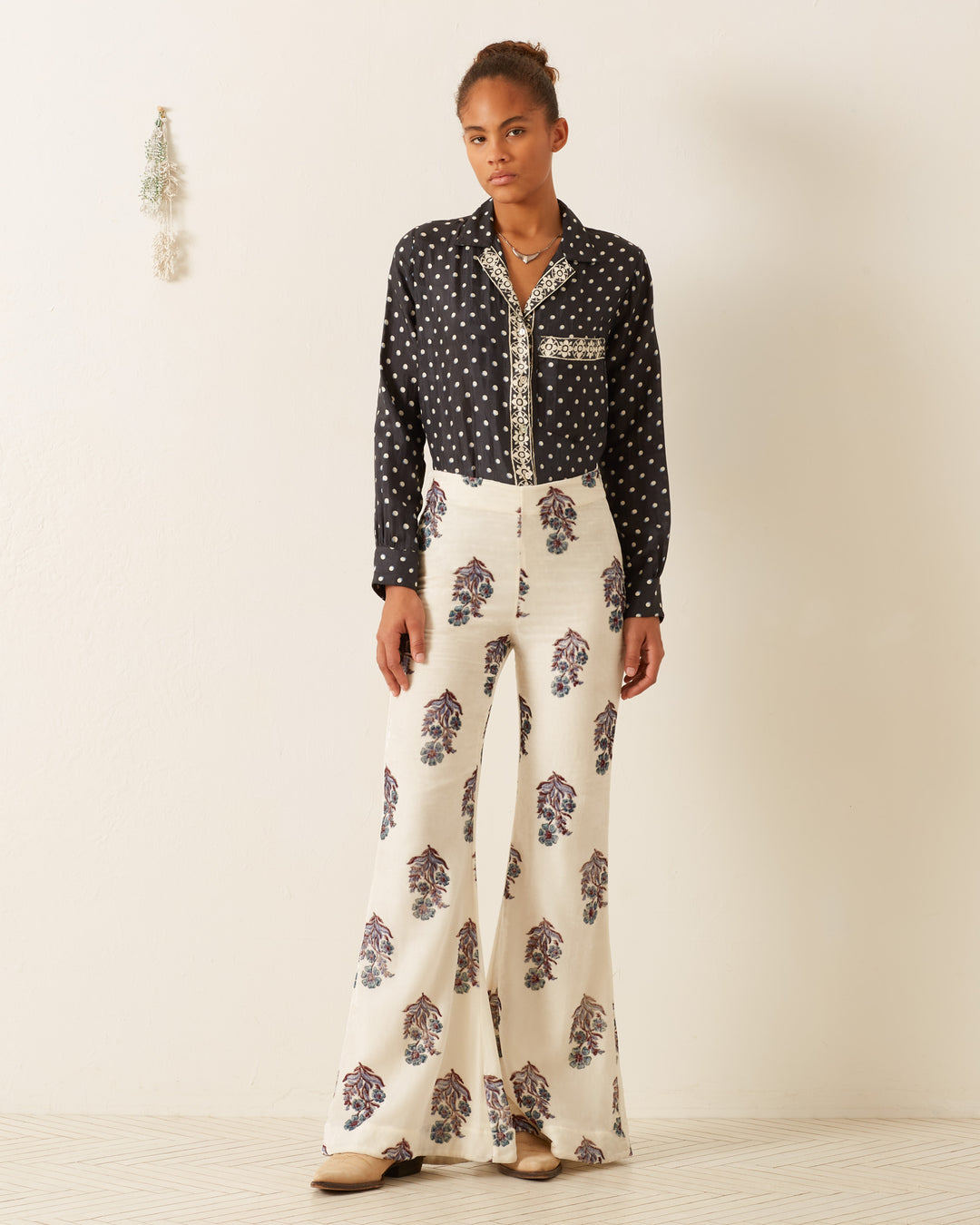 A person stands against a white wall, wearing the Patti Raven Dot Silk Shirt paired with white wide-legged pants featuring a floral print in a pajama-style cut. Their hair is tied up, and they complement the outfit with neutral-colored shoes. Decorative items hang on the wall in the background.