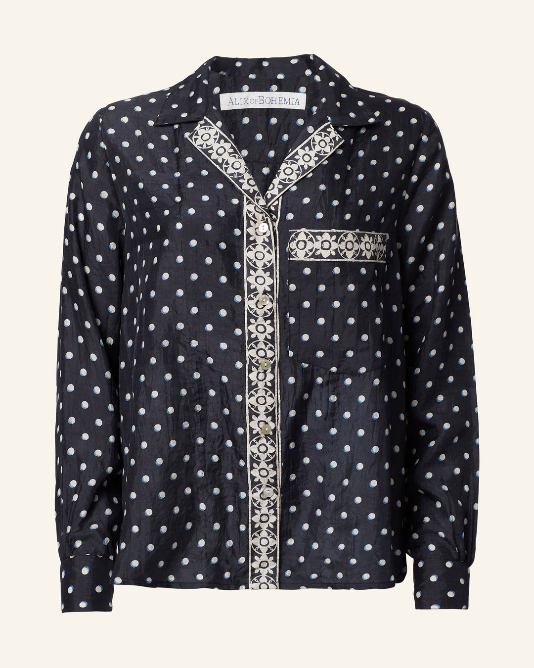 The Patti Raven Dot Silk Shirt is a black silk blouse with white polka dots, showcasing a pajama-style cut, a button-down front, and contrasting patterned trim along the buttons and pocket.