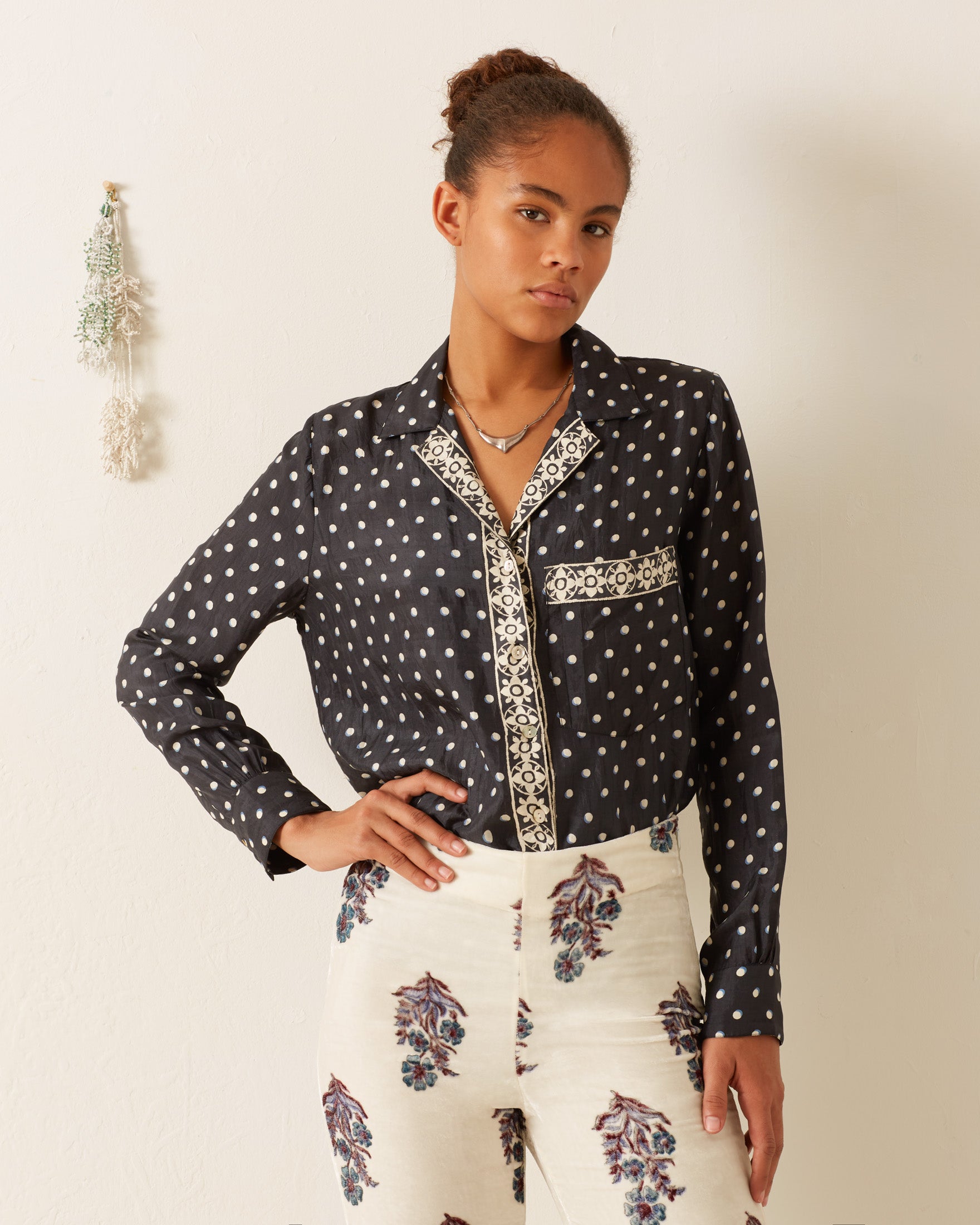 A person stands against a white wall wearing the Patti Raven Dot Silk Shirt, which features a black polka-dot pattern with a decorative trim, paired with light-colored pants showcasing a floral, hand-block printed design. A small decorative plant hangs on the wall, adding a touch of nature to the scene.