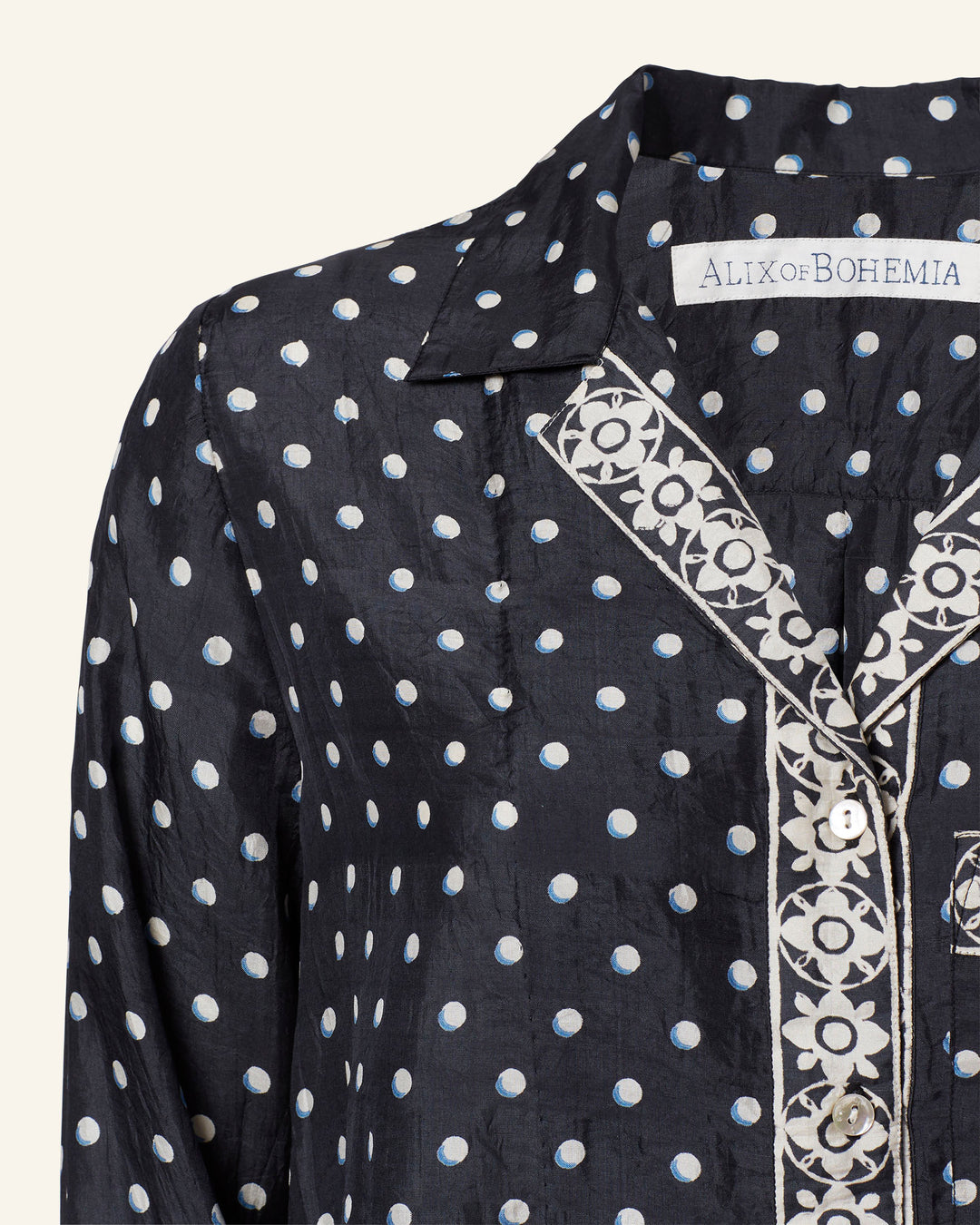 Close-up of a dark, pajama-style blouse adorned with white polka dots and intricate white embroidery near the collar and button seam. A label reading "ALIX of BOHEMIA" is attached inside the neckline, completing this chic Patti Raven Dot Silk Shirt.